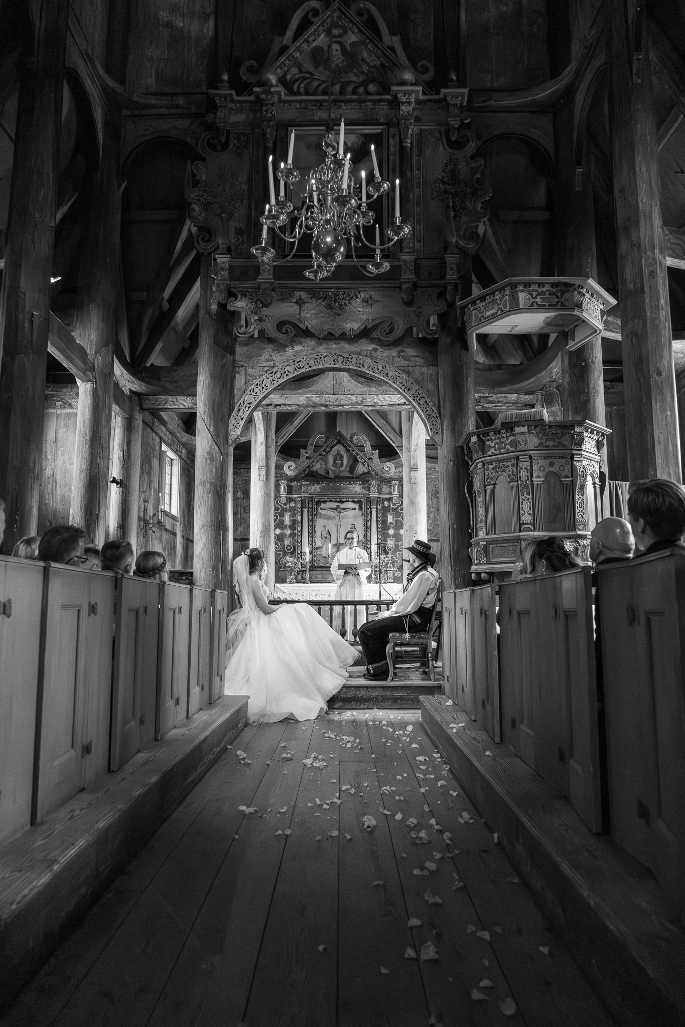 Black and white photo of bride and groom seated at the altar of an ornate historic wooden church during ceremony.