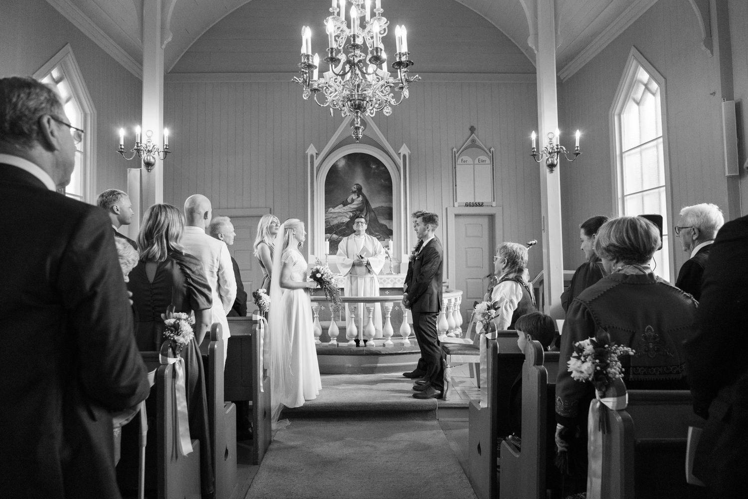 Bride and groom exchange vows at altar in small white church with guests seated in pews.