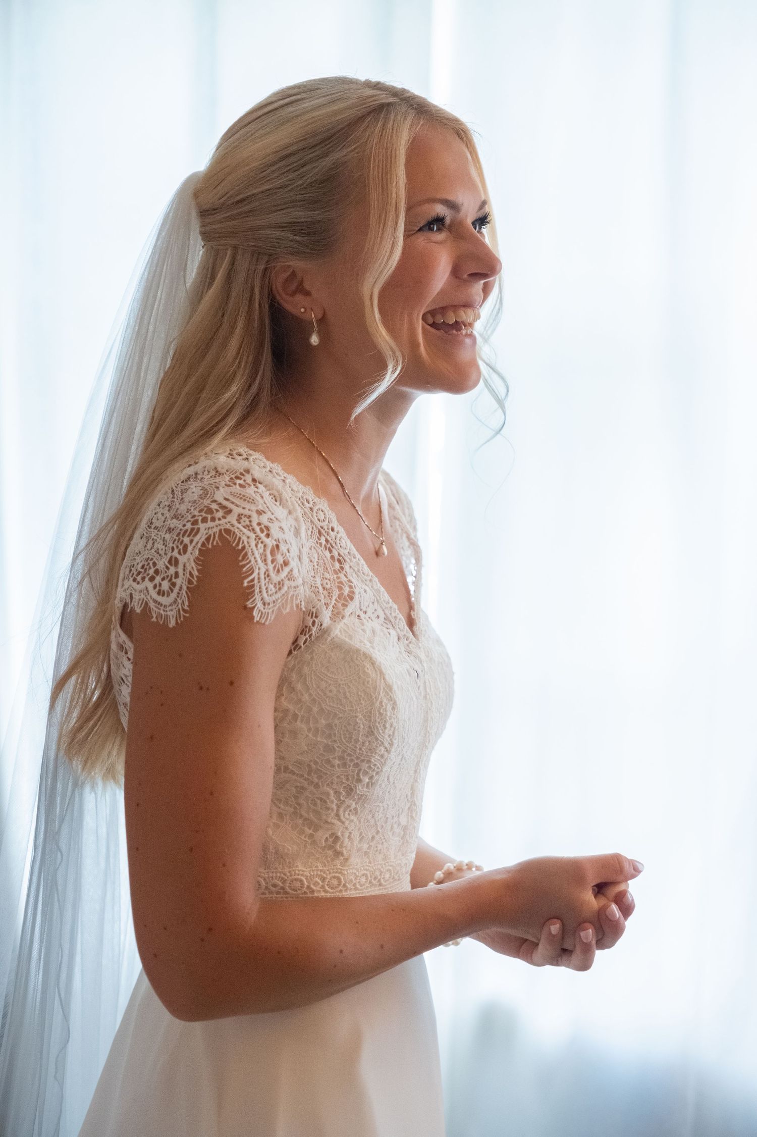 Smiling blonde bride in lace wedding dress and veil standing by window on wedding day.