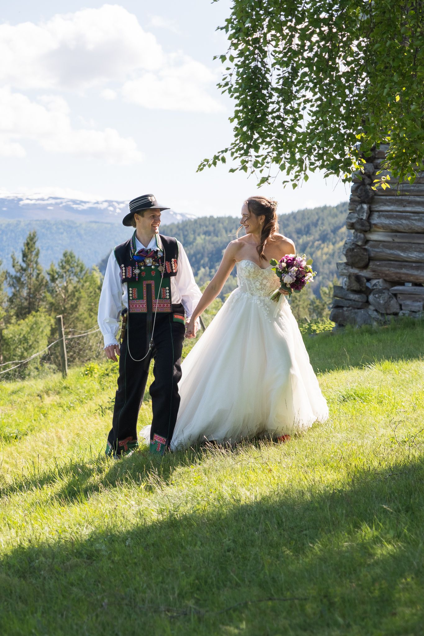 Bride and groom in Norwegian bunad walking hand in hand on green hillside with mountains and rustic building nearby.