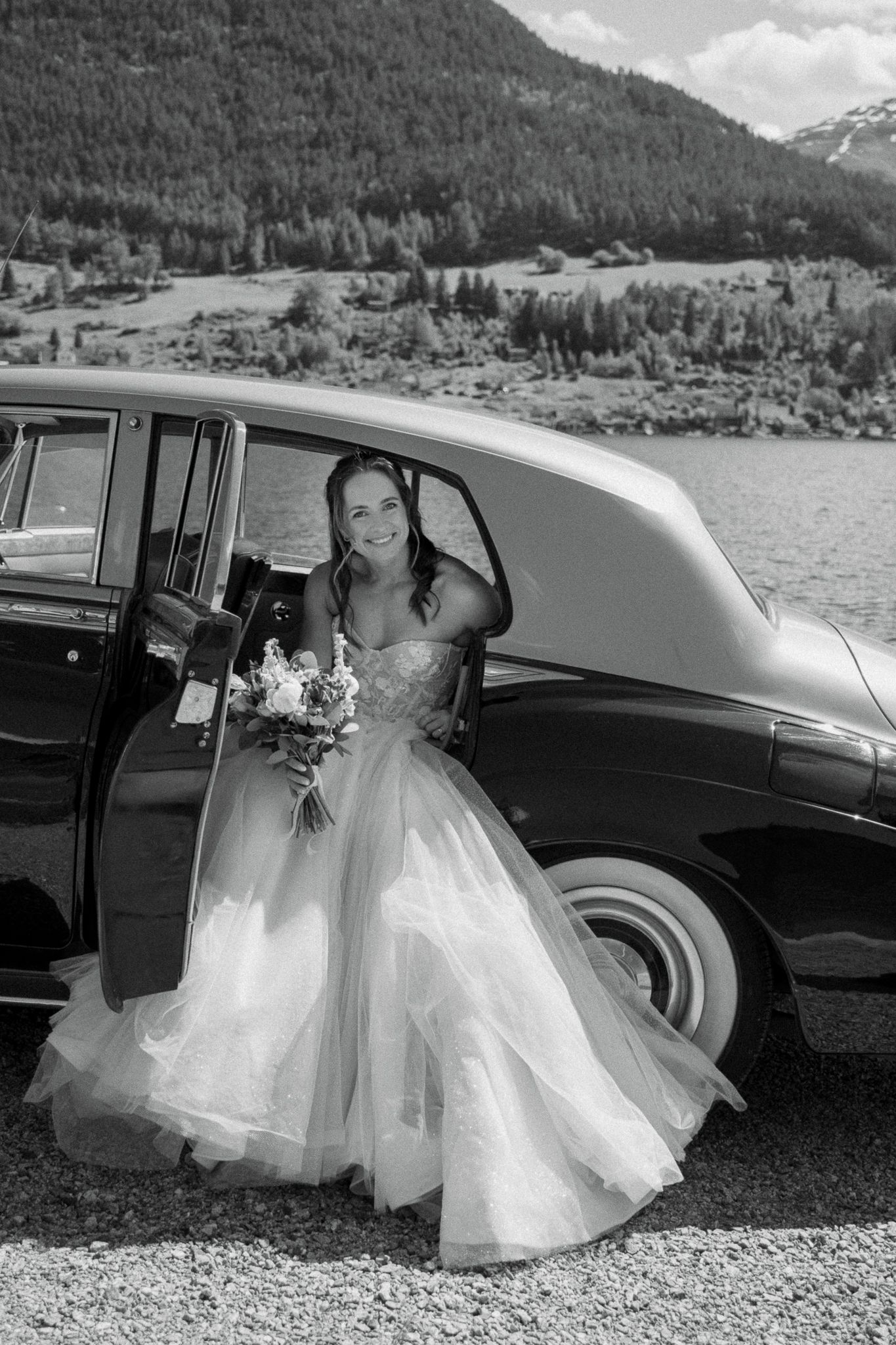 Smiling bride in strapless gown holds bouquet while posing beside a vintage classic car lakeside.