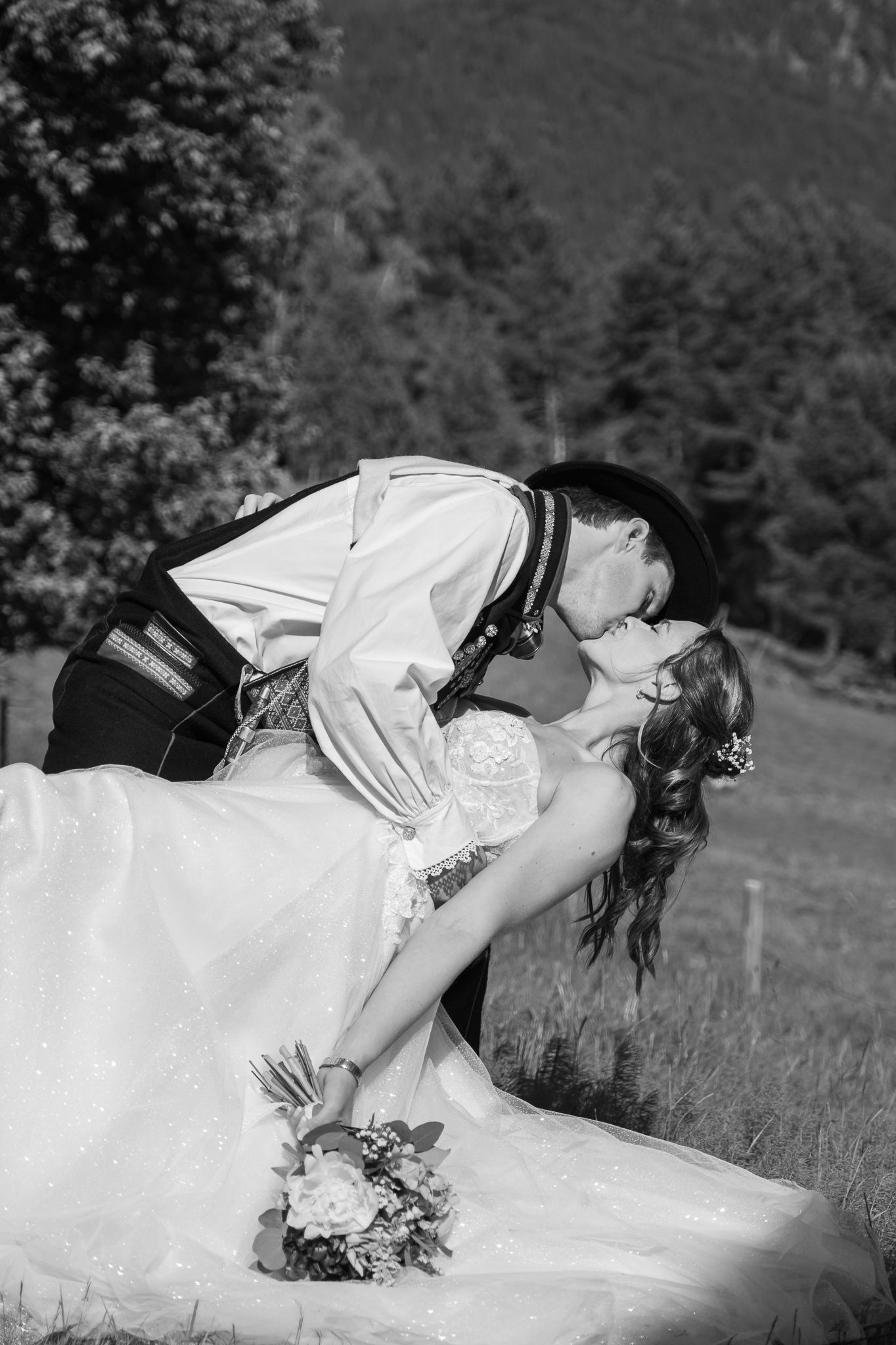 Black and white romantic photo of groom dipping and kissing bride on a grassy hillside with forest backdrop.