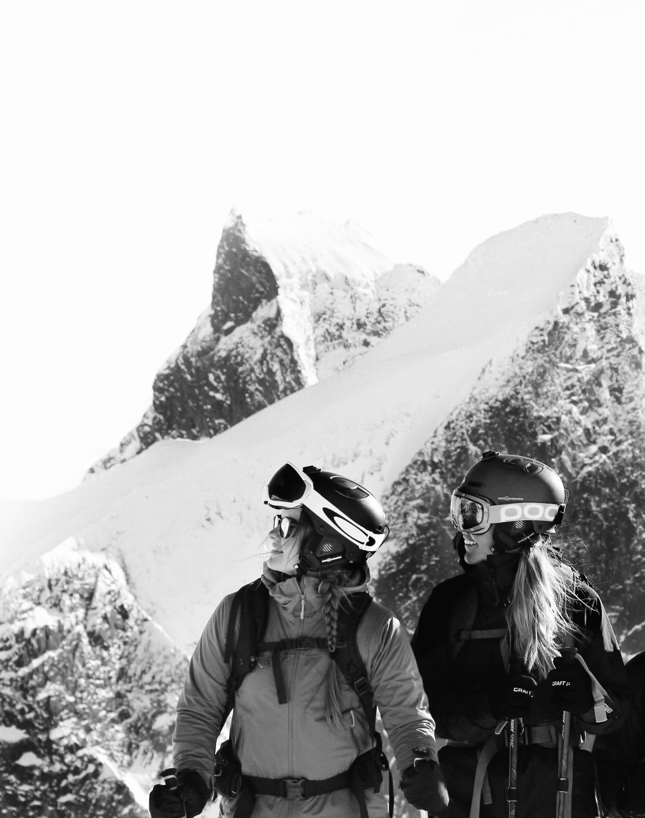 Black and white photo of two skiers with helmets and goggles standing before a towering snow-covered mountain peak.