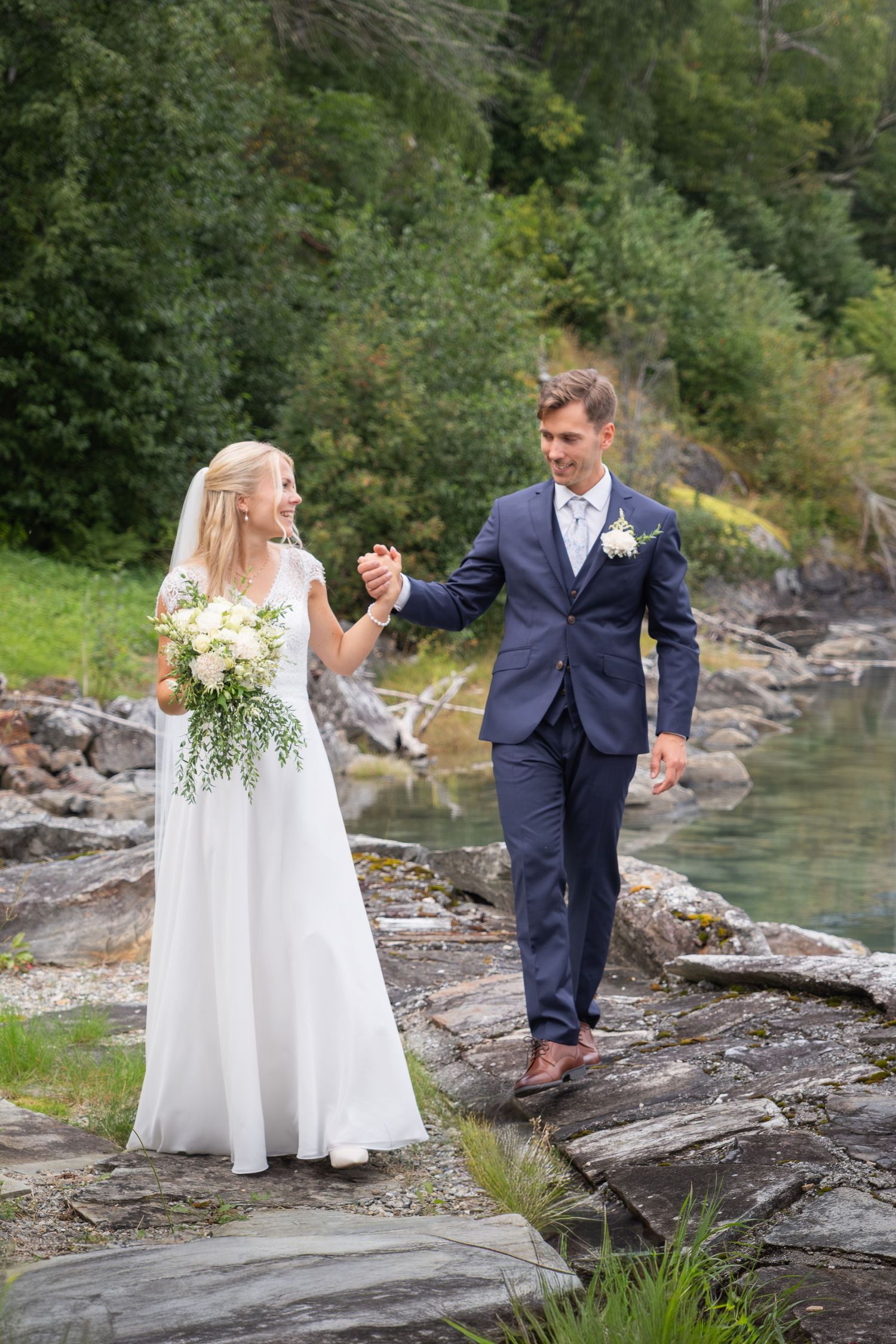 Bride and groom walk hand in hand along rocky riverside path in lush green nature.