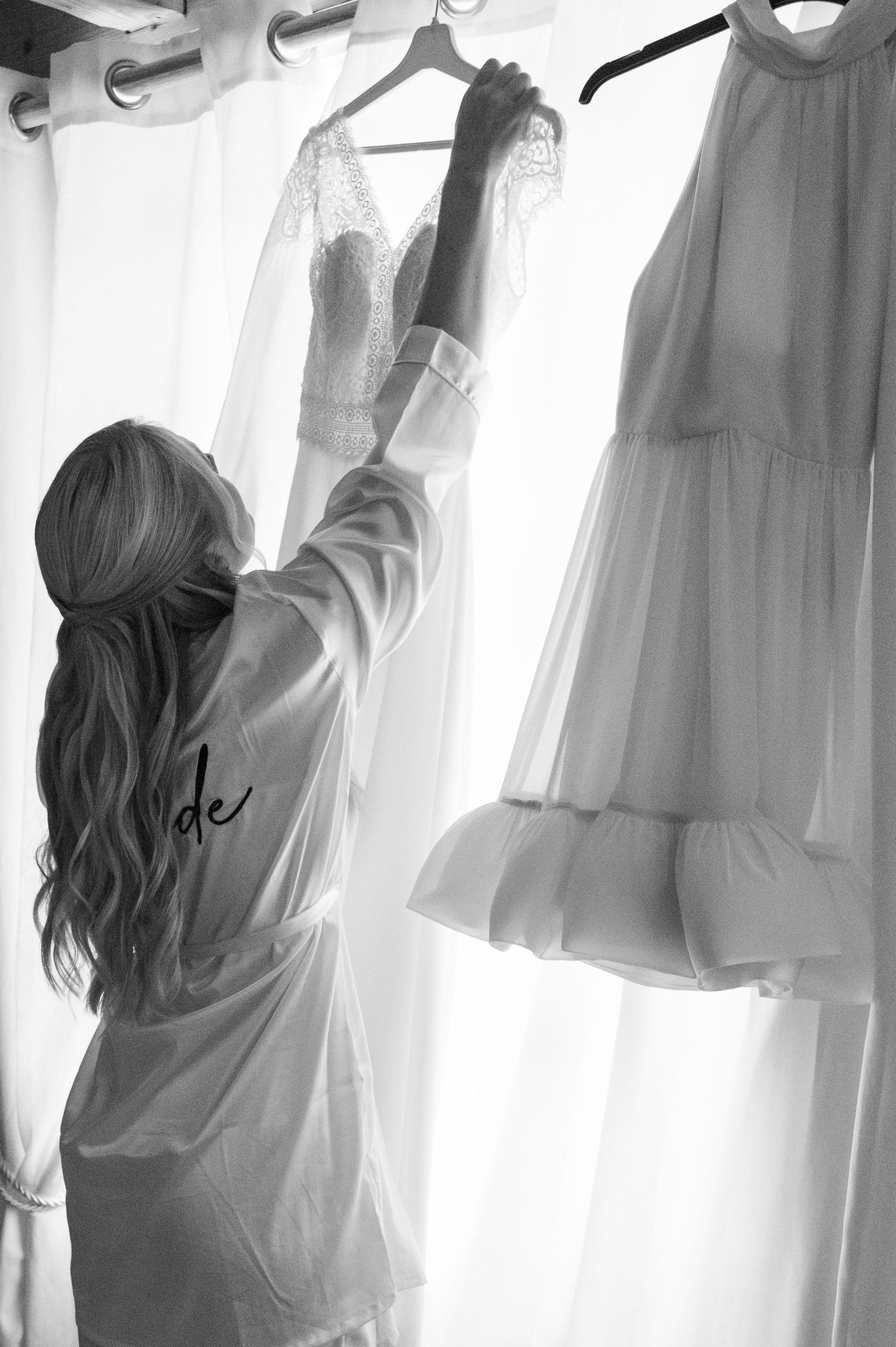 Black and white photo of bride in robe reaching for her lace wedding dress on a hanger.