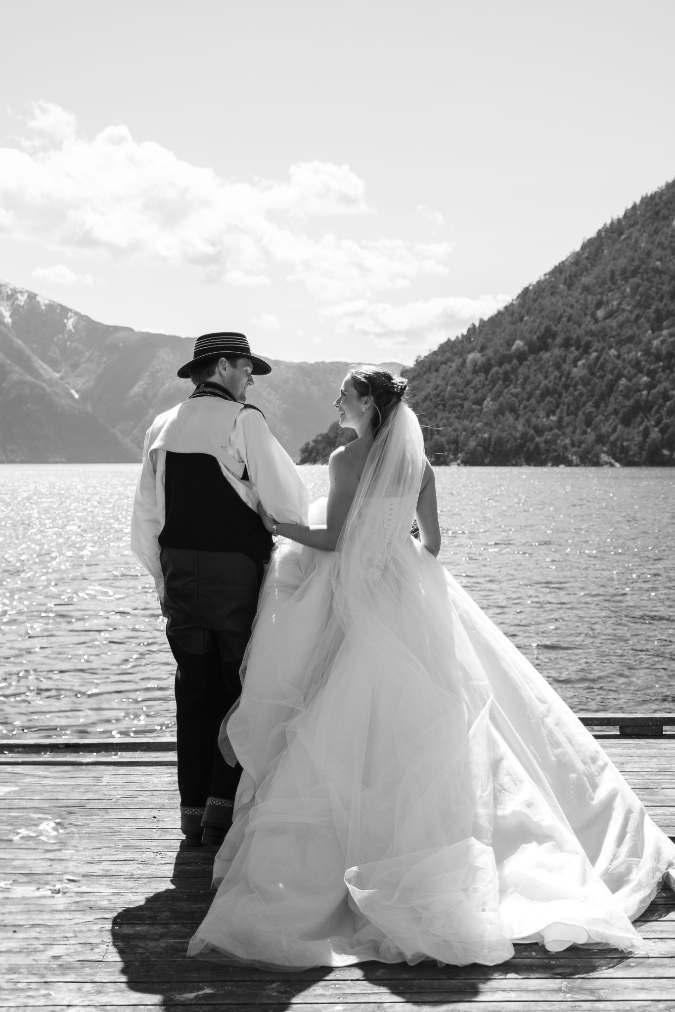Black and white wedding portrait of couple standing on dock gazing at scenic mountain lake view.