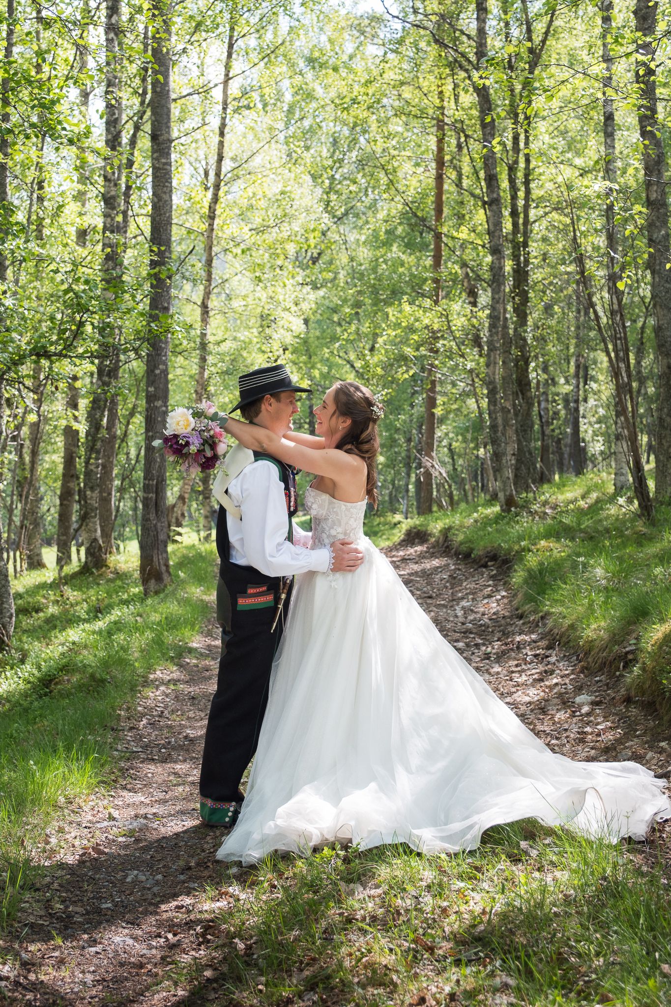 Bride and groom embracing on forest path surrounded by tall green trees in outdoor wedding portrait.