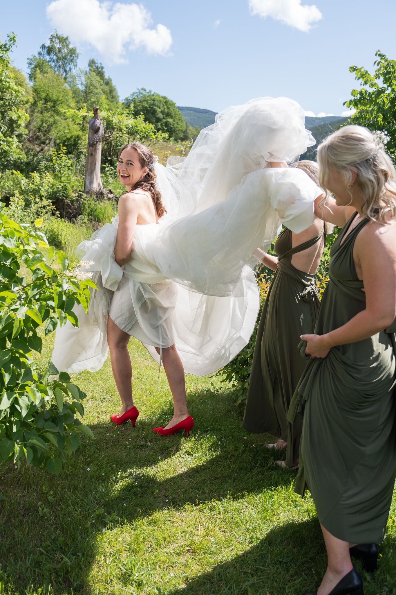 Laughing bride in red shoes has white gown lifted by bridesmaids in green dresses outdoors.