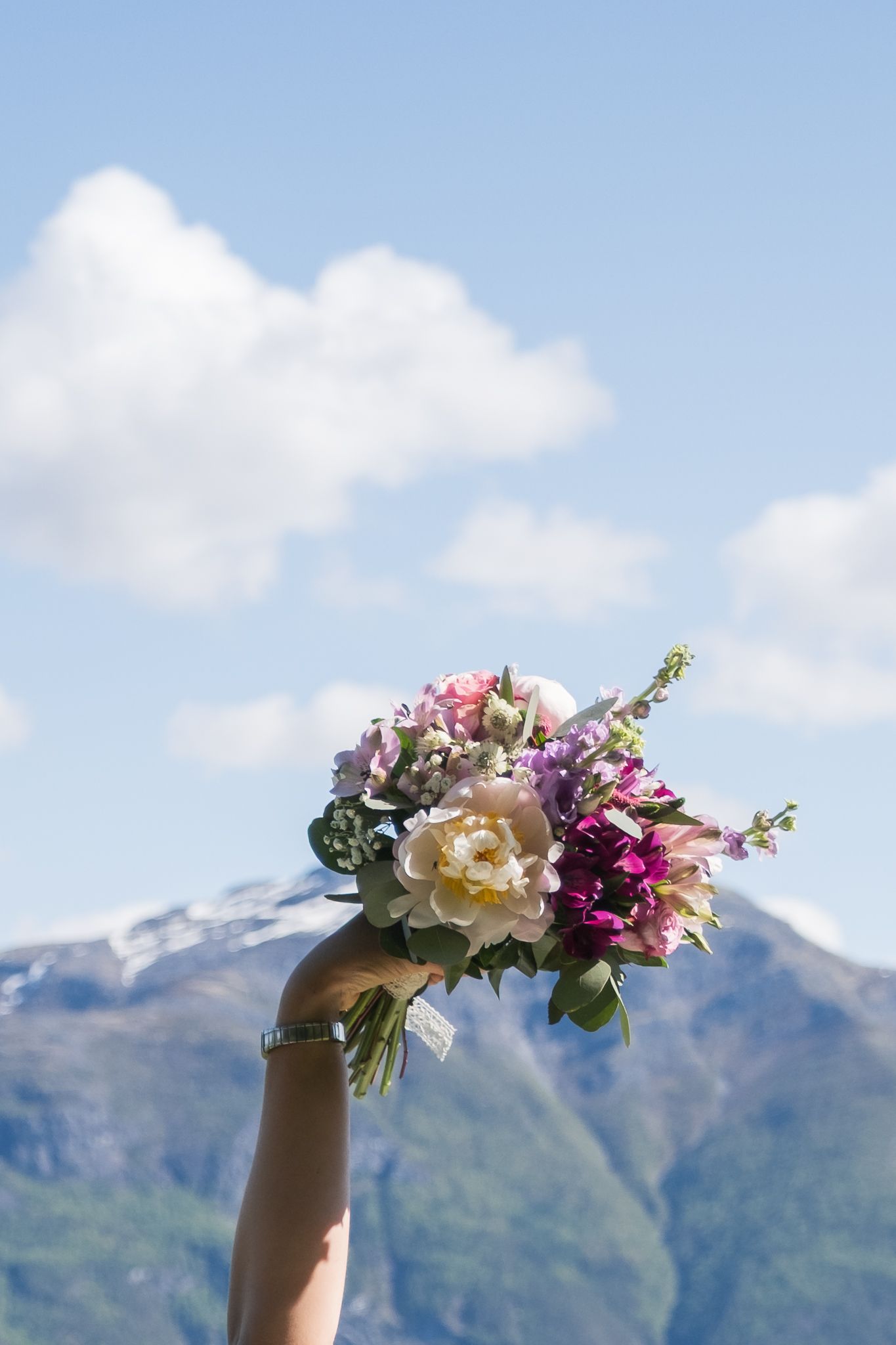 Bride holding a colorful wedding bouquet of peonies and roses high against a blue mountain sky.