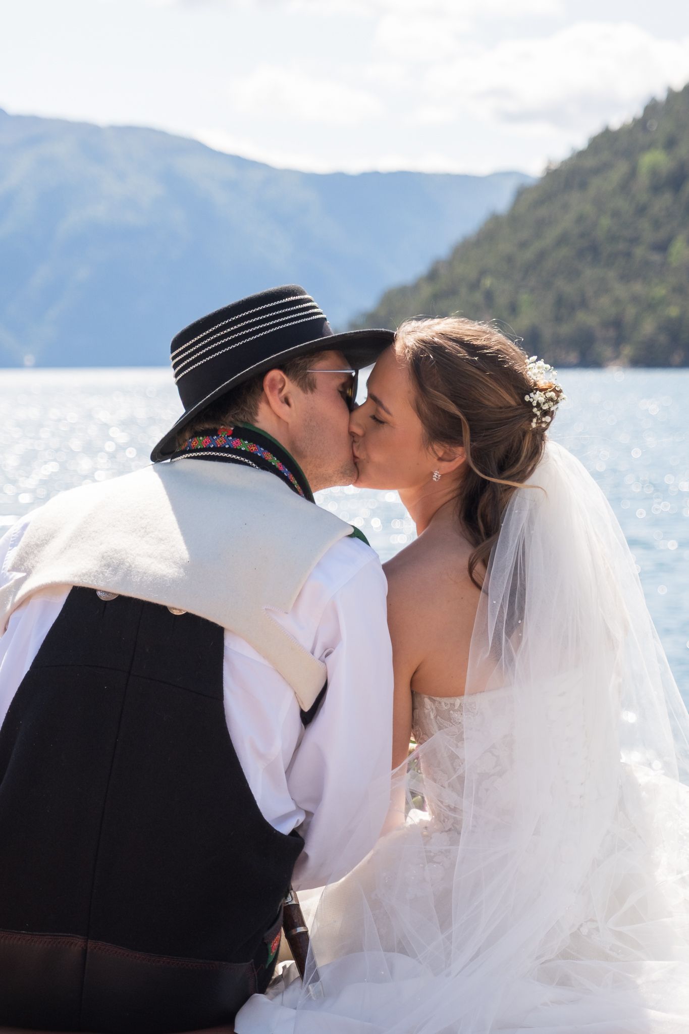 Bride and groom sharing a kiss while seated by a sparkling mountain fjord on their wedding day.