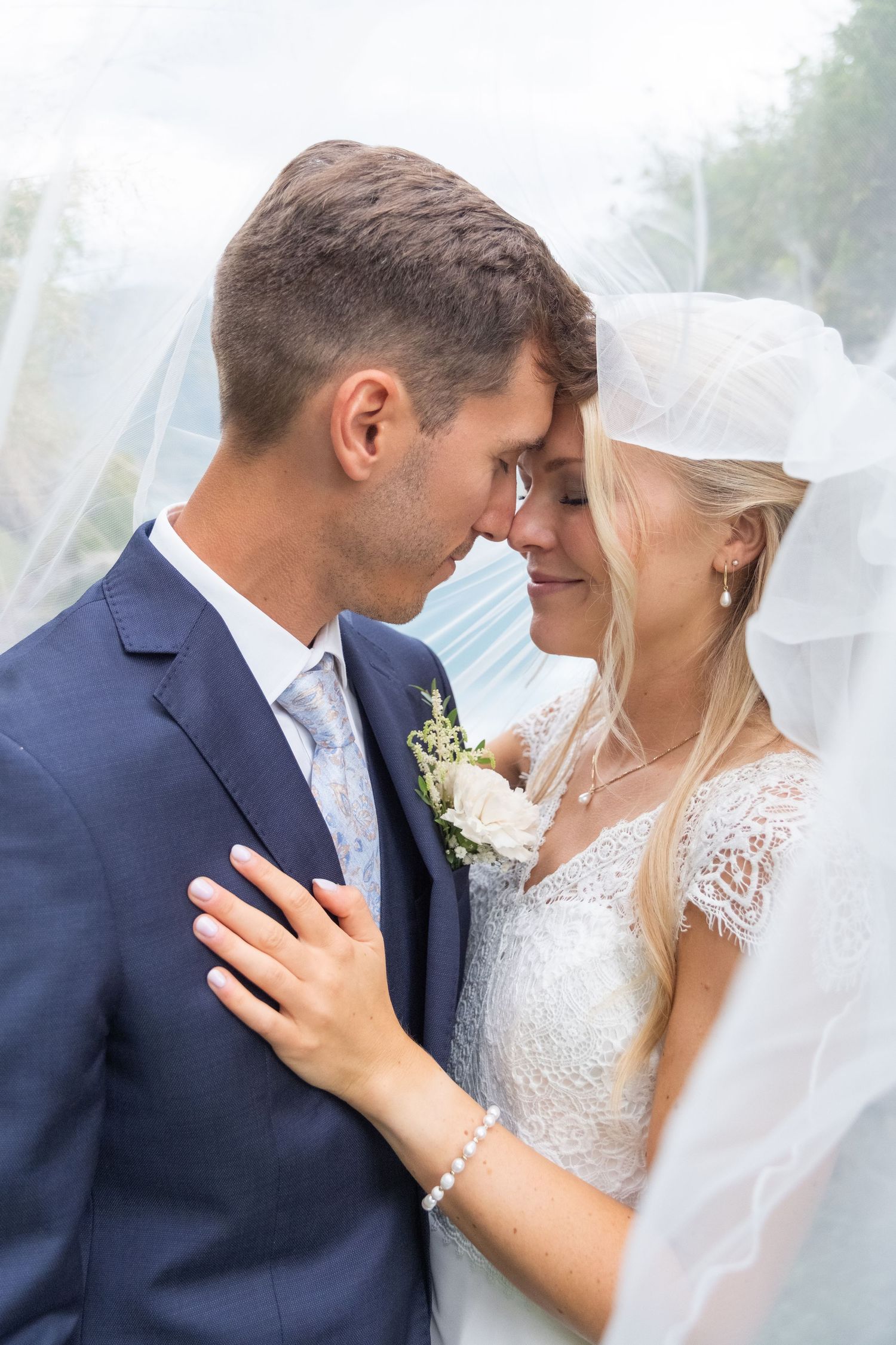 Bride and groom share an intimate forehead-to-forehead moment beneath the wedding veil outdoors.