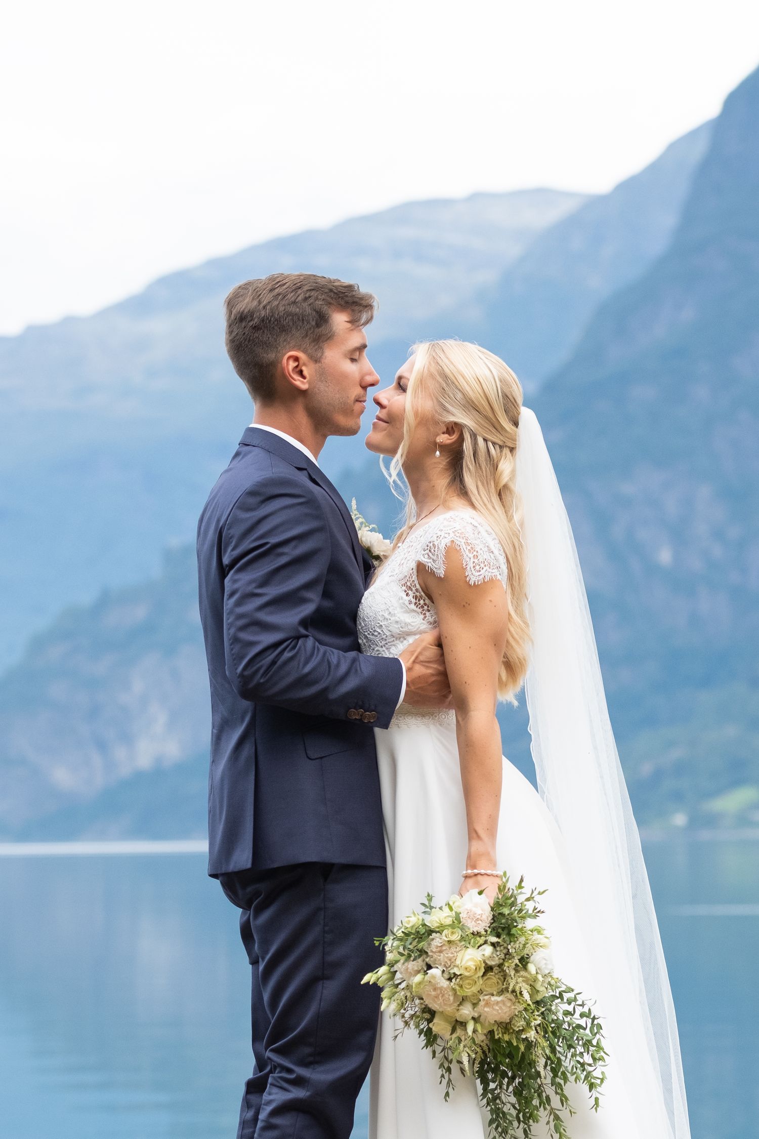 Groom in navy suit embraces blonde bride holding bouquet near a stunning mountain lake wedding venue.
