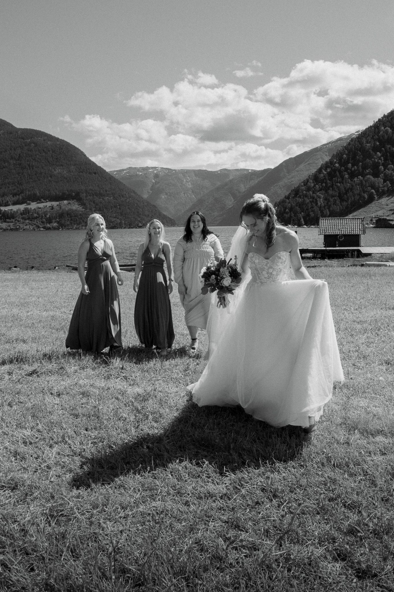 Bride holding bouquet walks across grass while three bridesmaids in green dresses watch by a mountain lake.