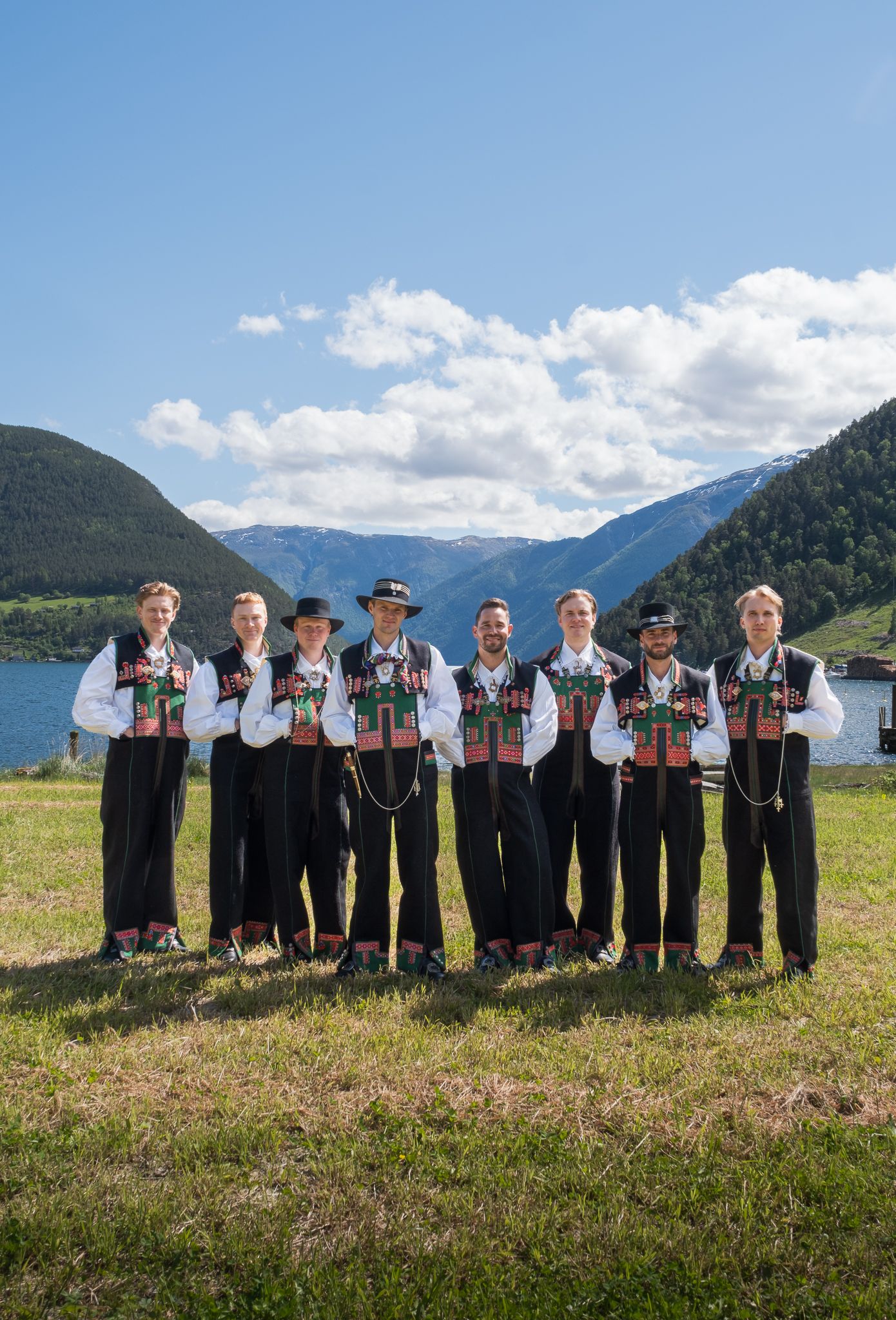 Seven men in traditional Norwegian bunad folk costumes standing together by a mountain fjord on a sunny day.
