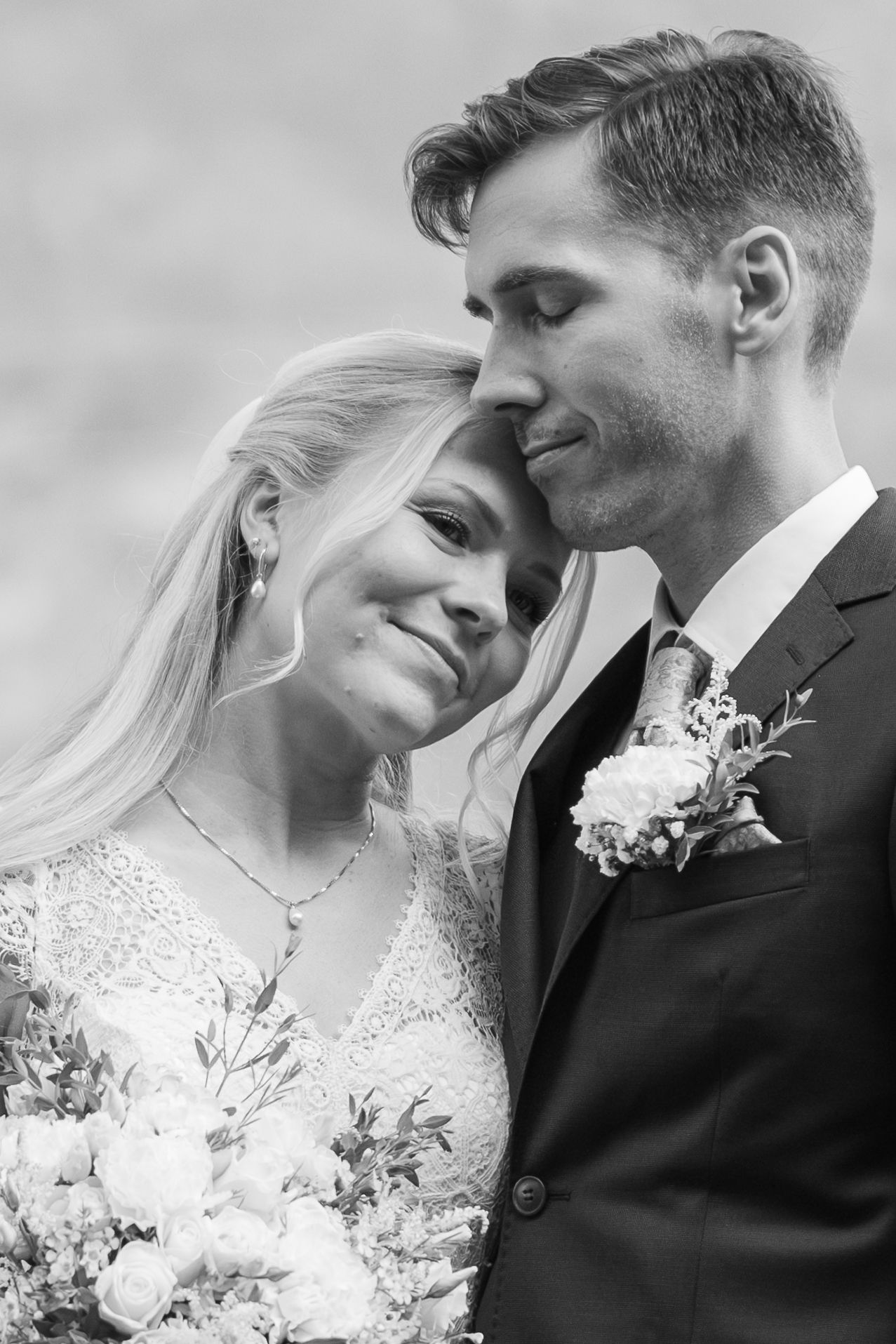 Bride leans head on grooms shoulder as he holds her close in intimate black and white portrait.