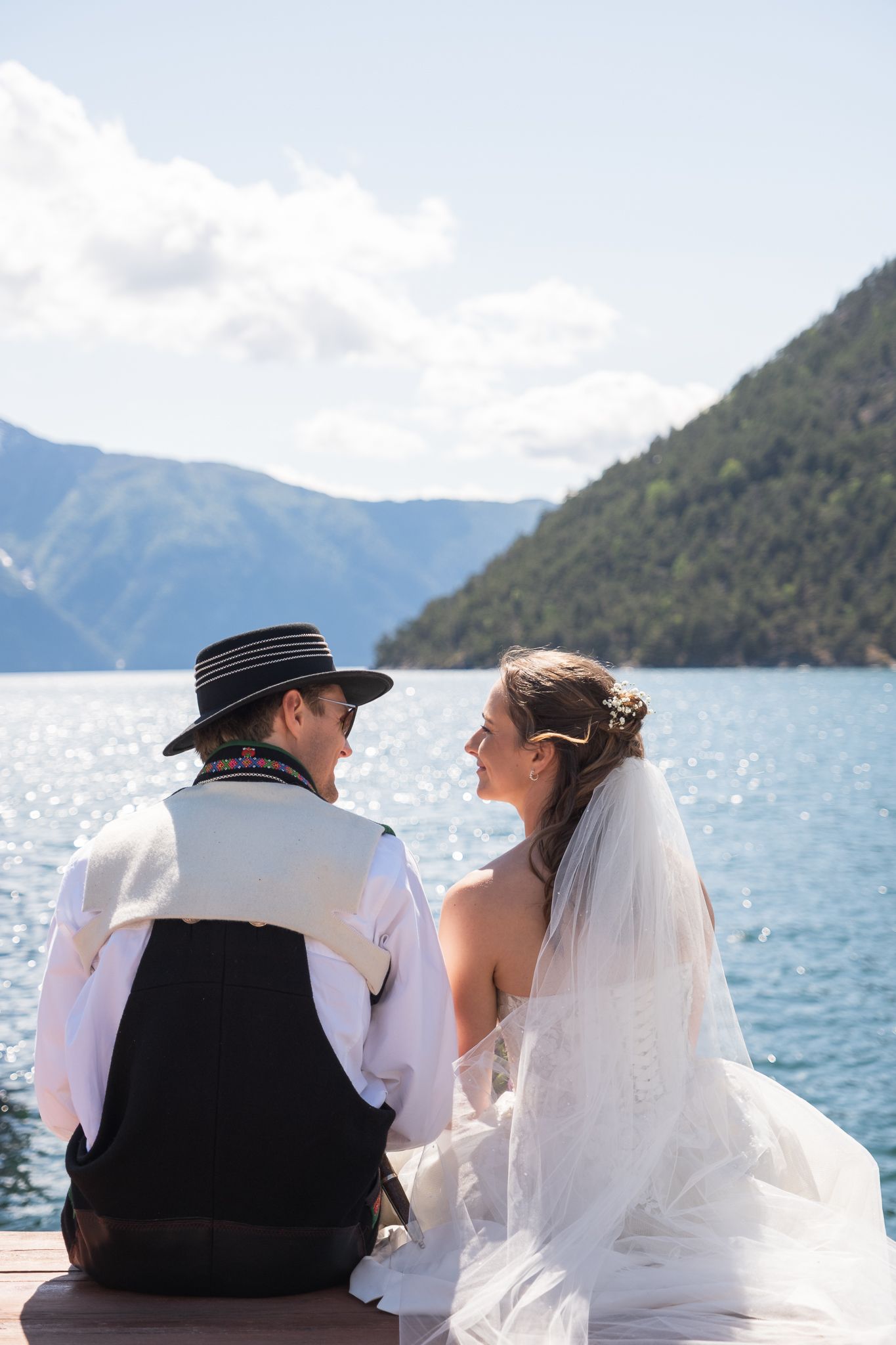 Bride and groom sit on dock sharing intimate moment with scenic mountain lake behind them.