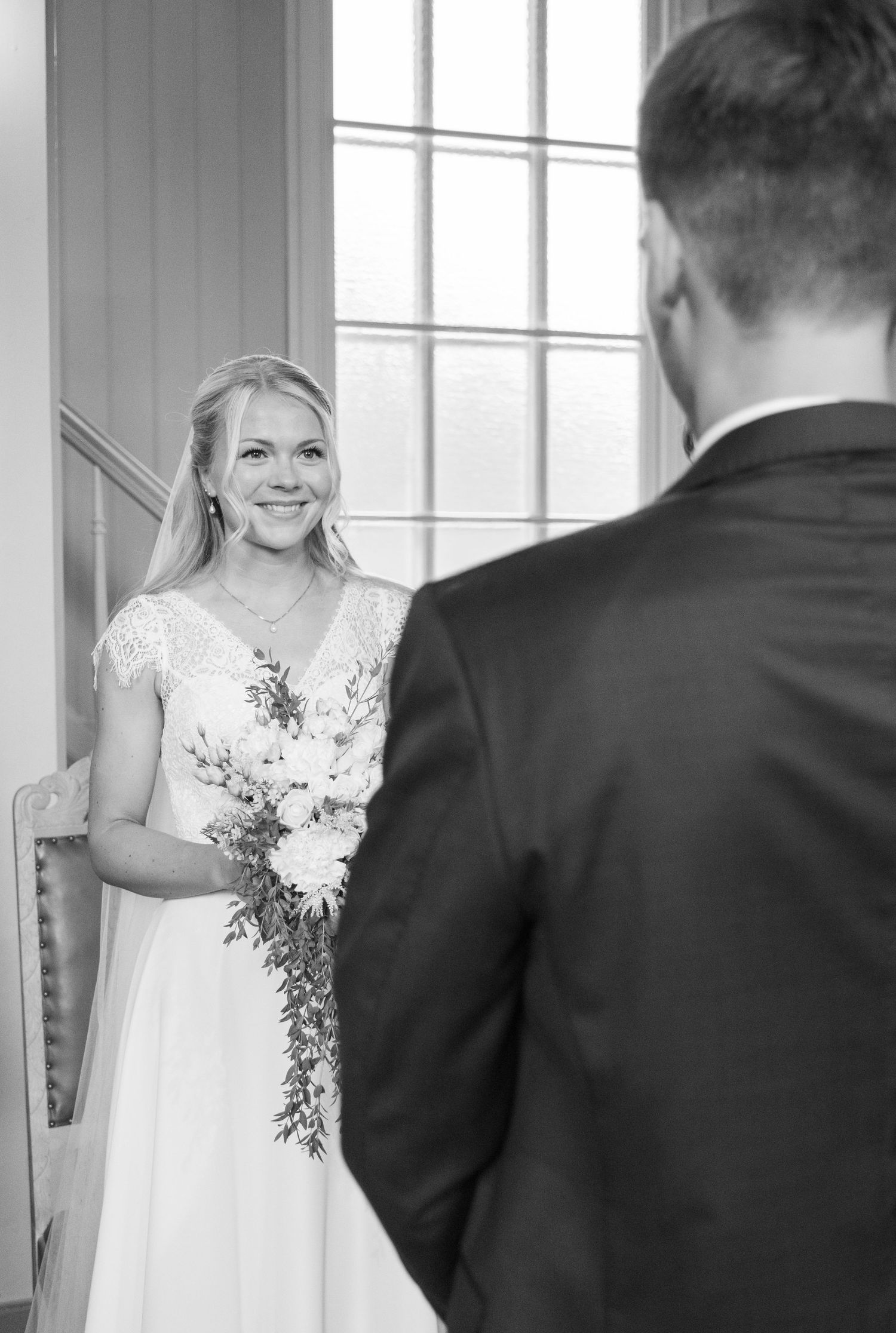 Bride in lace wedding gown smiles at groom during first look moment in black and white photo.