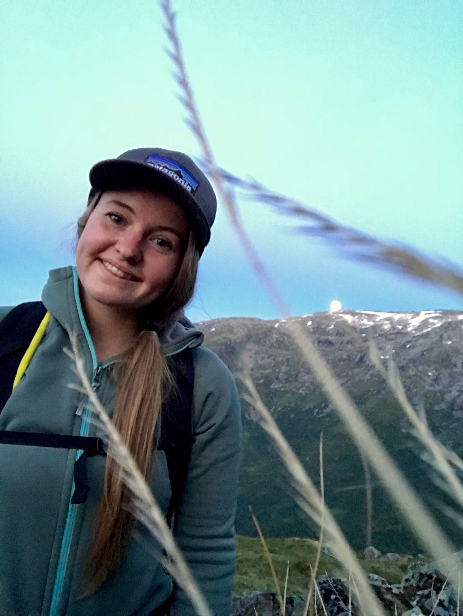 Happy young woman in a Patagonia cap smiling with mountain scenery behind her at twilight.