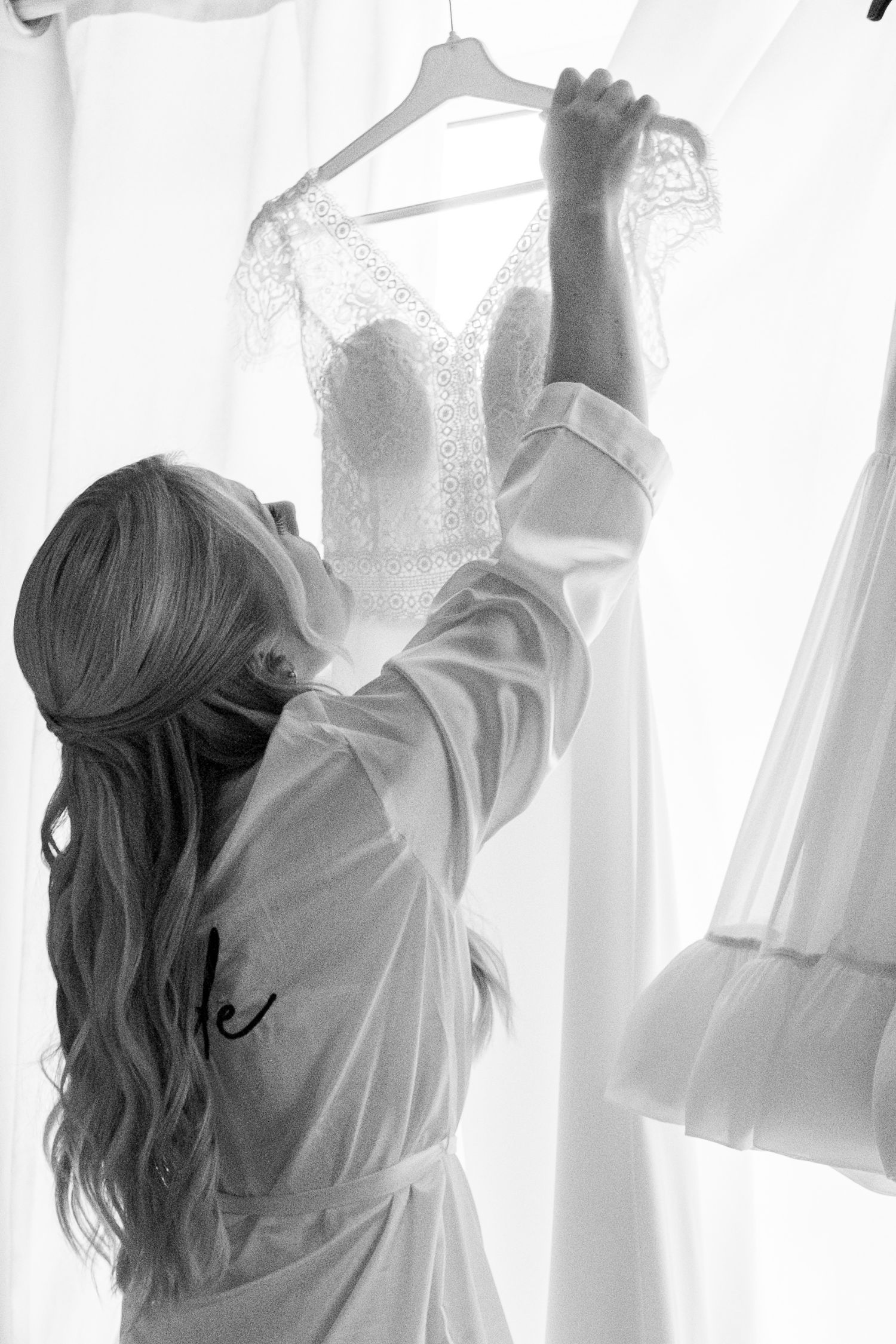 Bride in a robe reaching up to touch her lace wedding dress on a hanger in black and white.