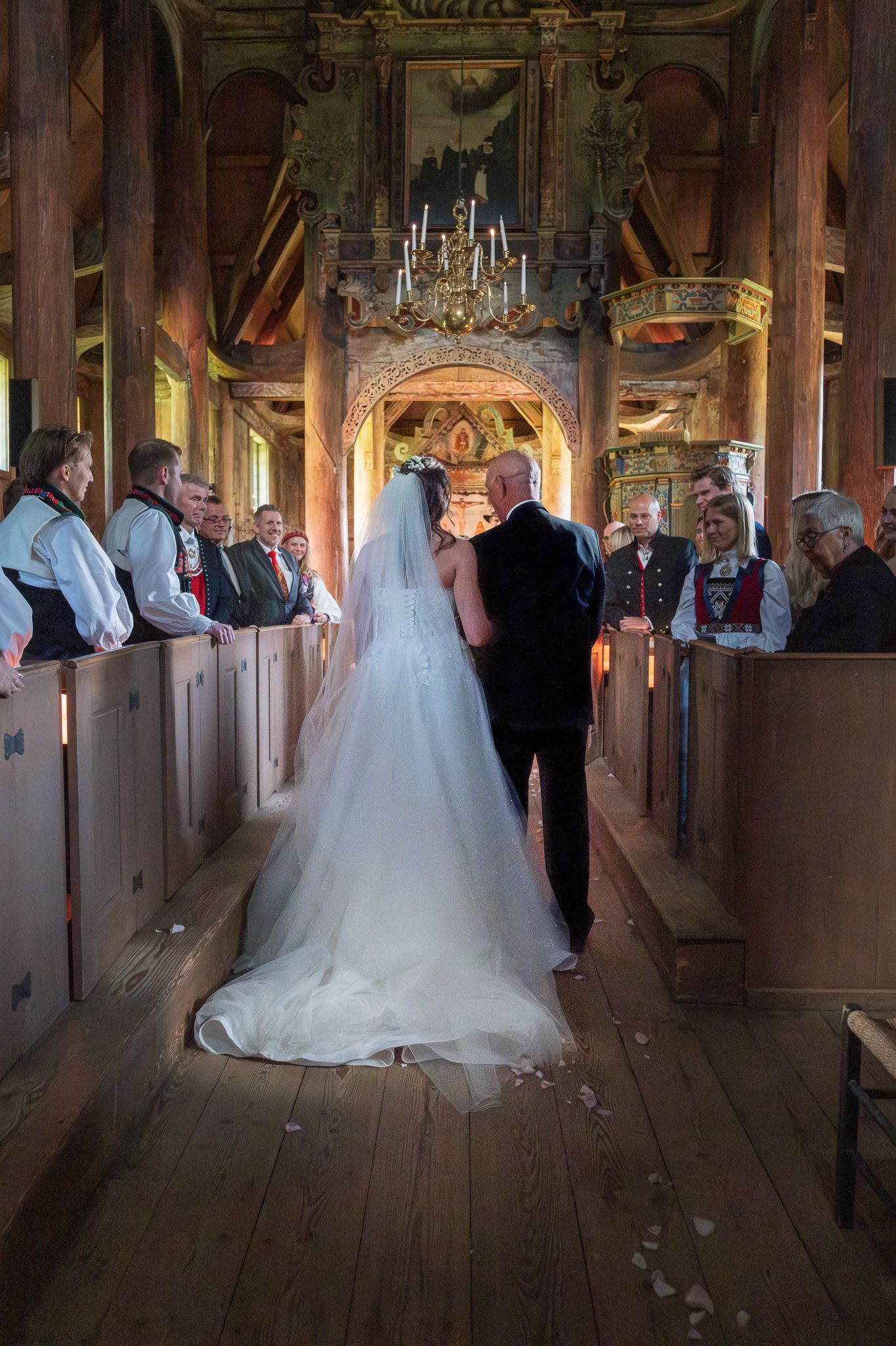 Bride and escort walk down the aisle of a historic wooden church with ornate decor and wedding guests watching.