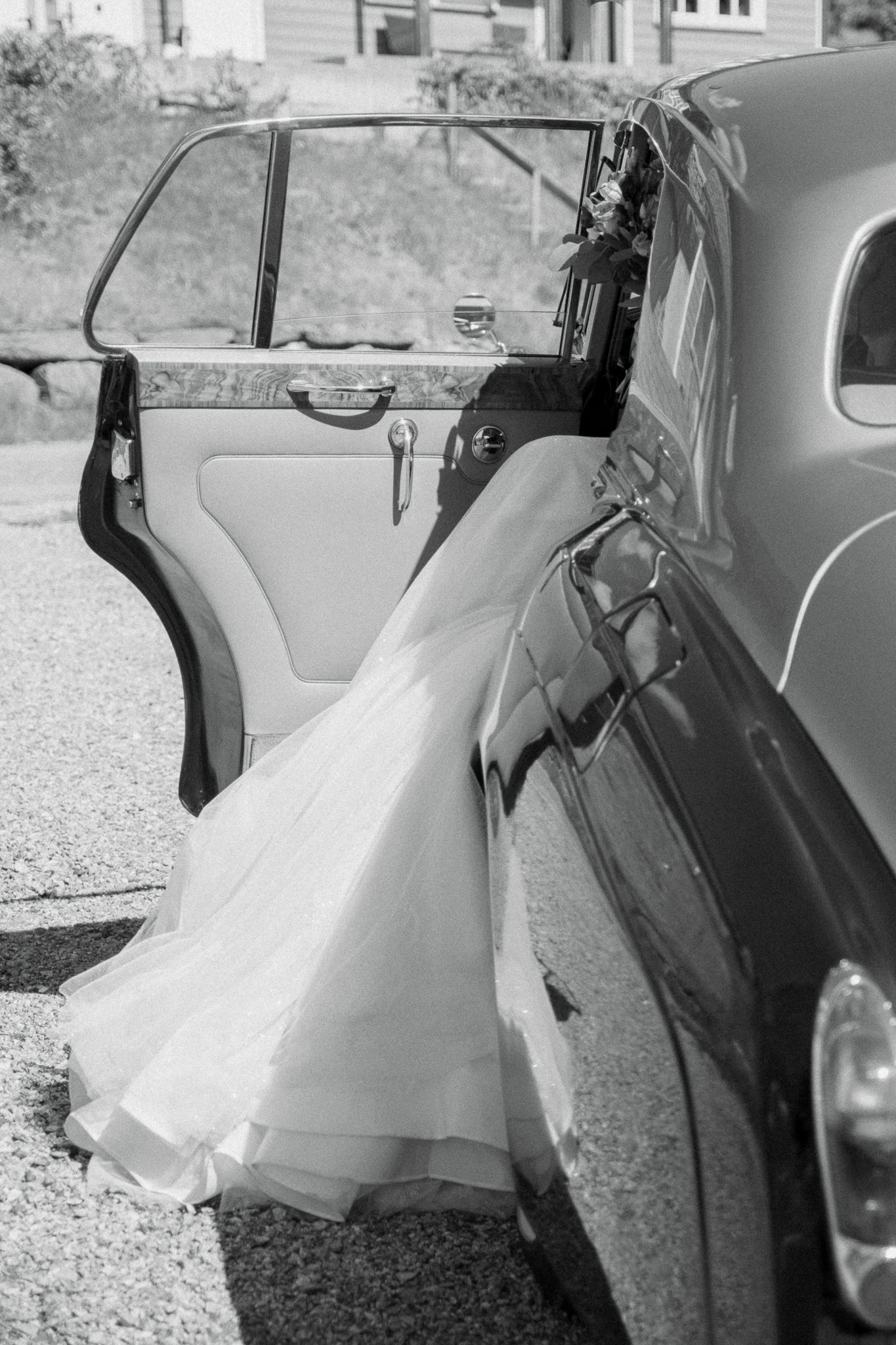 Black and white photo of a bride's white wedding gown flowing out of a vintage classic car door.