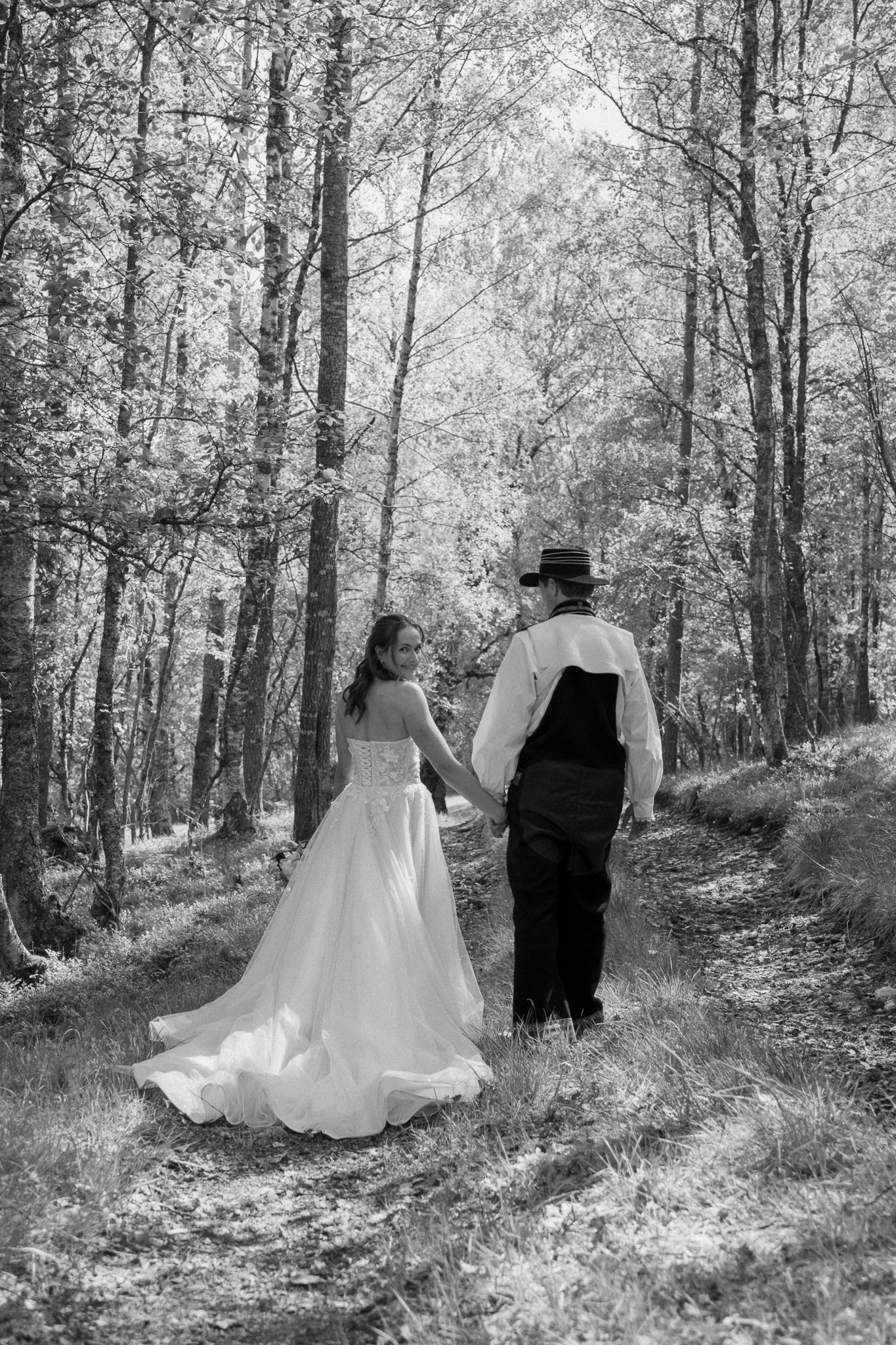 Black and white photo of a bride and groom walking hand in hand through a forest path.