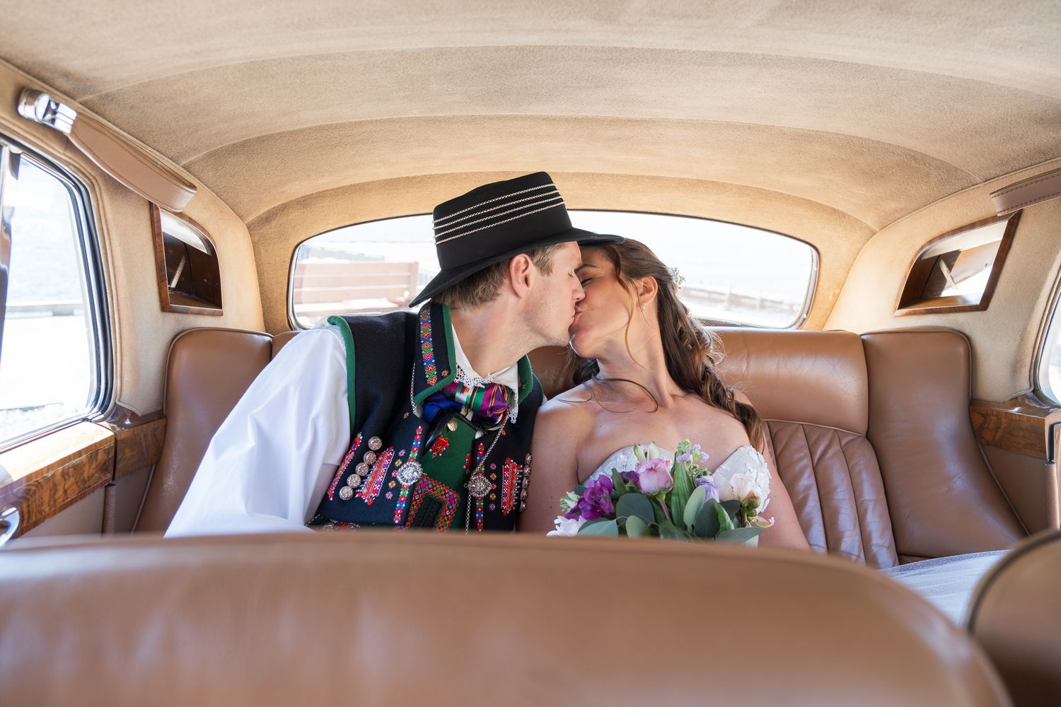 Bride and groom share a kiss in the backseat of a vintage classic car on their wedding day.
