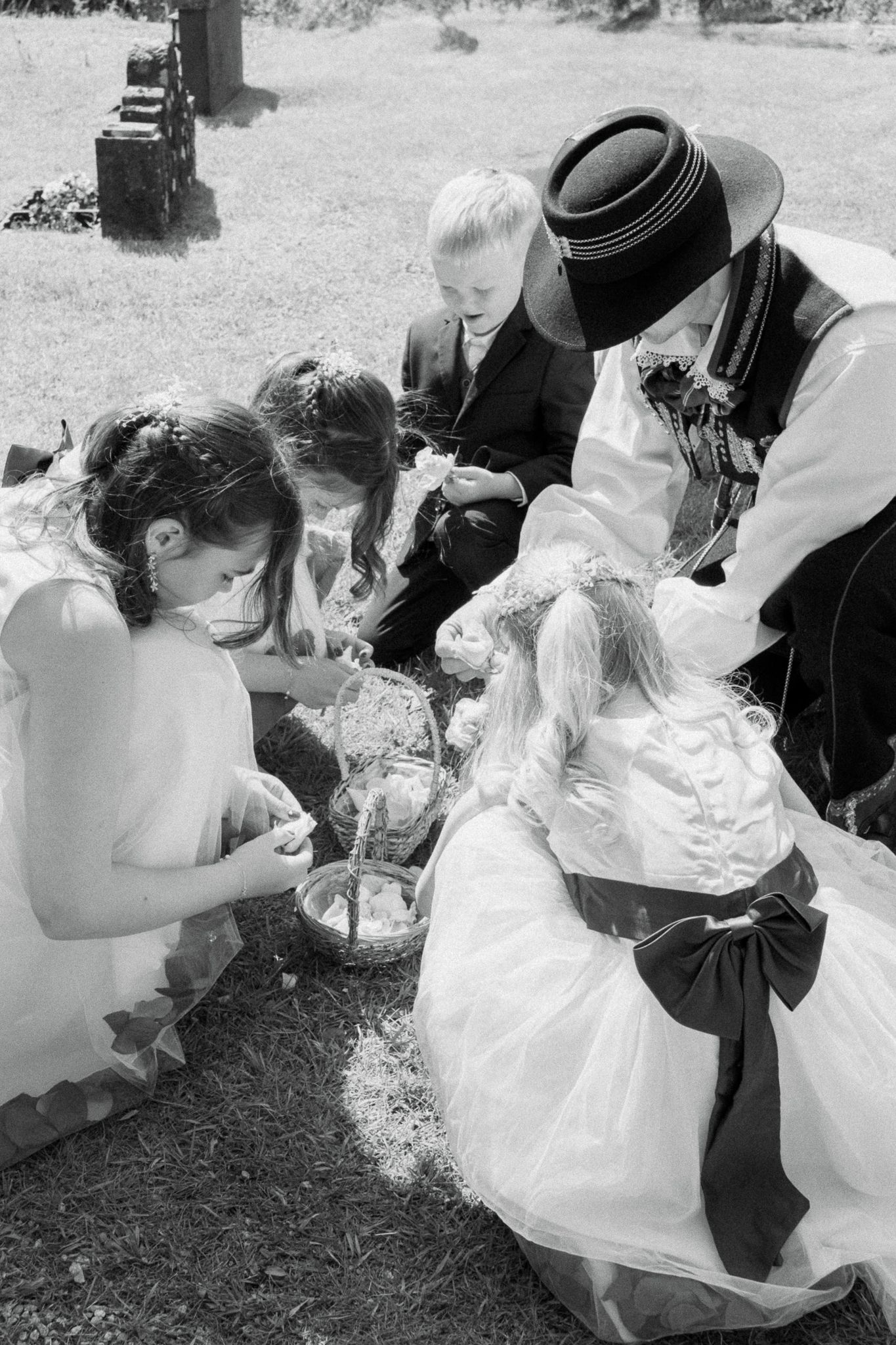 Black and white photo of wedding party children and adult in traditional attire gathered on grass examining a basket.