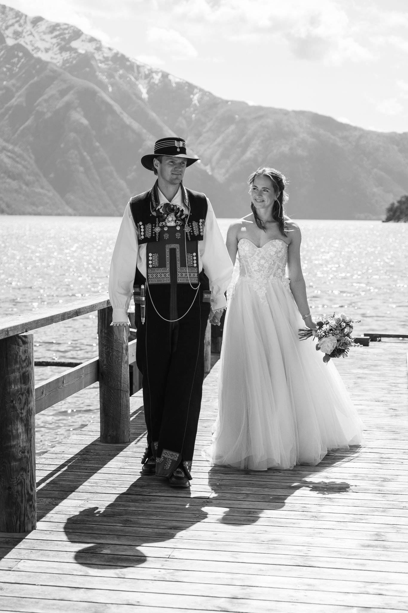 Bride and groom walking hand in hand on a wooden pier, groom in traditional Norwegian bunad costume.