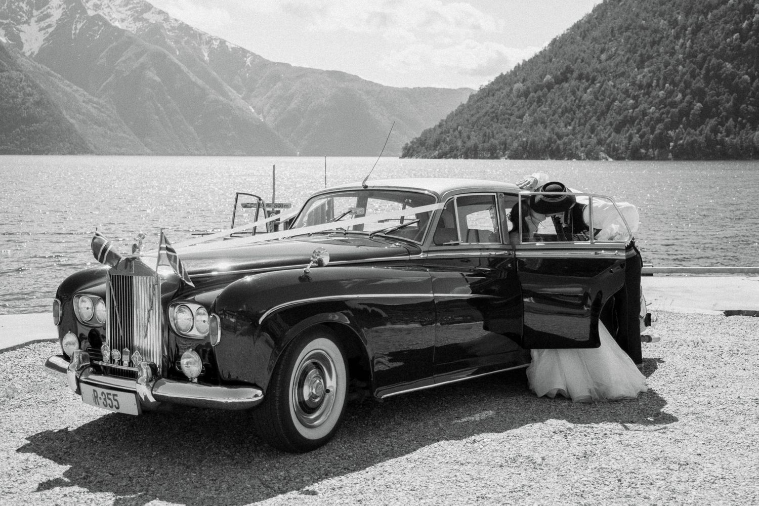 Black and white photo of a classic vintage Rolls-Royce wedding car parked beside a mountain lake.