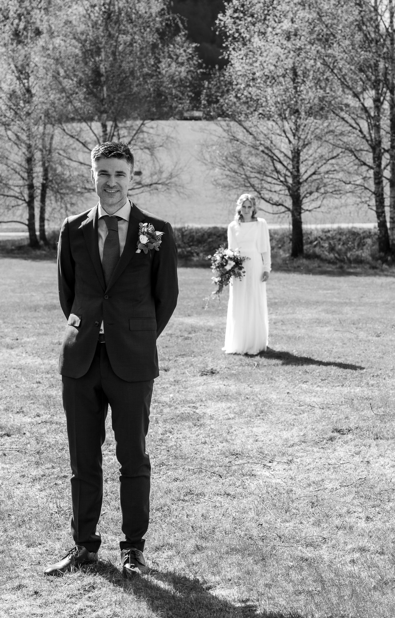 Groom standing in foreground with bride holding bouquet in background in a black and white outdoor wedding photo.