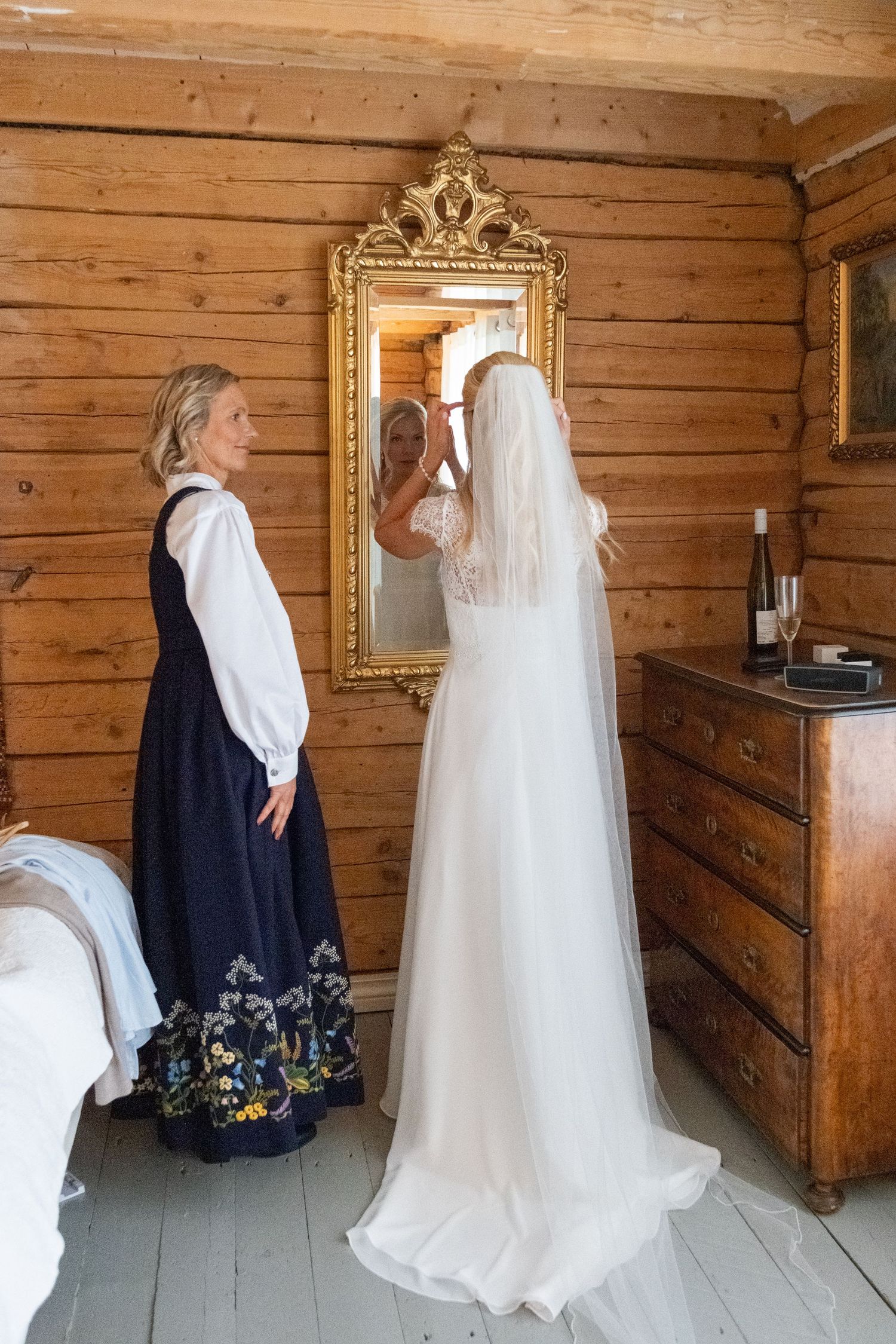 Bride in white gown adjusting her veil in a gold ornate mirror in a rustic wooden room.