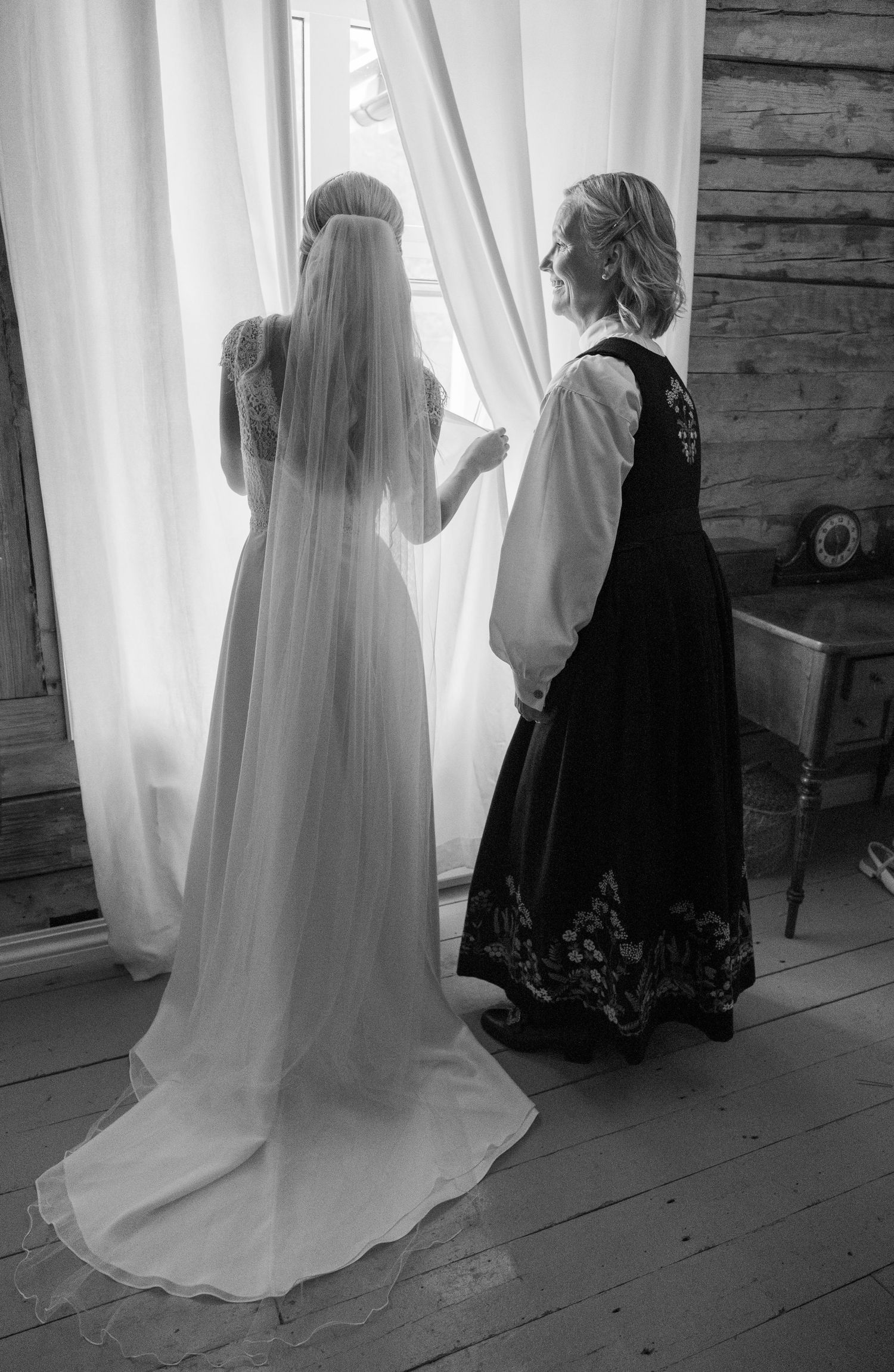 Bride in white wedding gown and veil standing at window with woman in traditional Norwegian bunad dress.