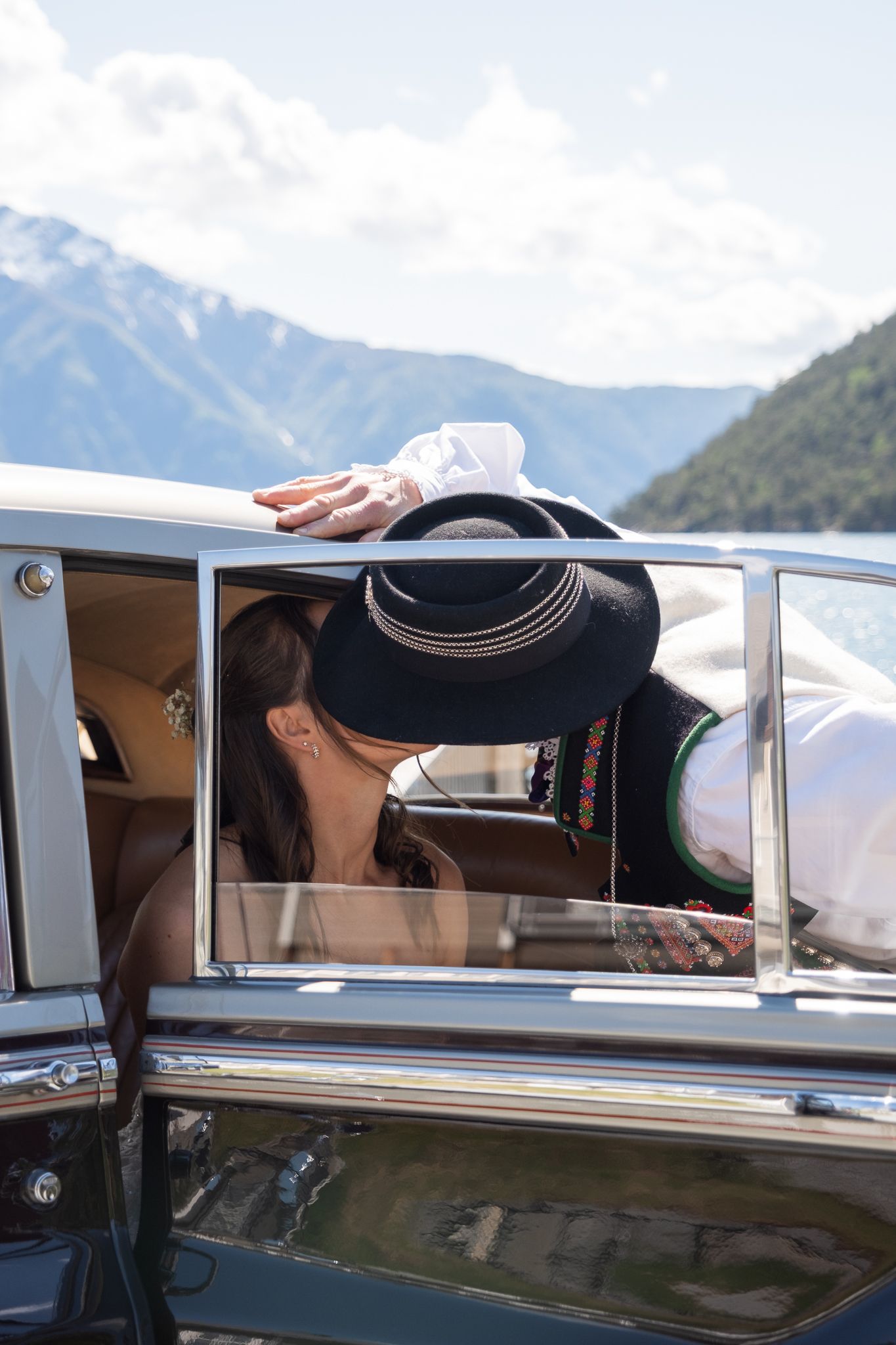 Bride and groom share a kiss through the window of a vintage classic car with mountain scenery.