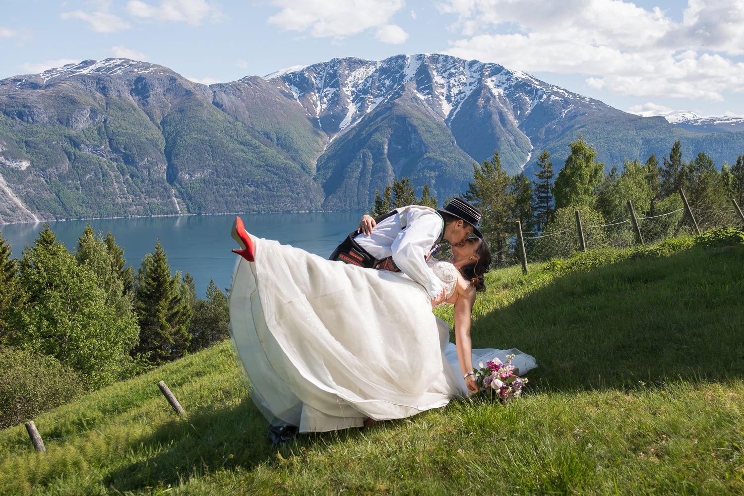 Groom dipping bride in a romantic kiss on a green hillside with stunning fjord and snow-capped mountains in background.
