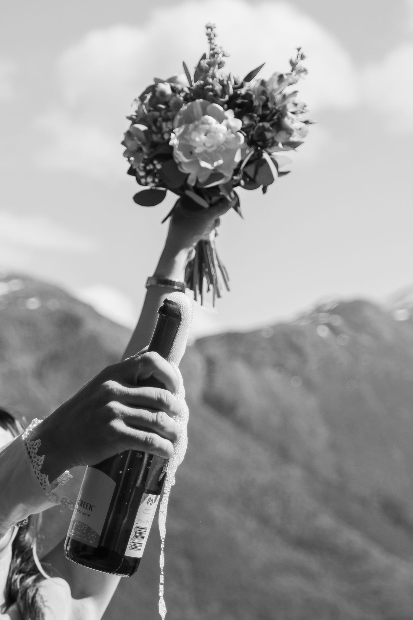 Bride raises flower bouquet and champagne bottle triumphantly against mountain backdrop in black and white.