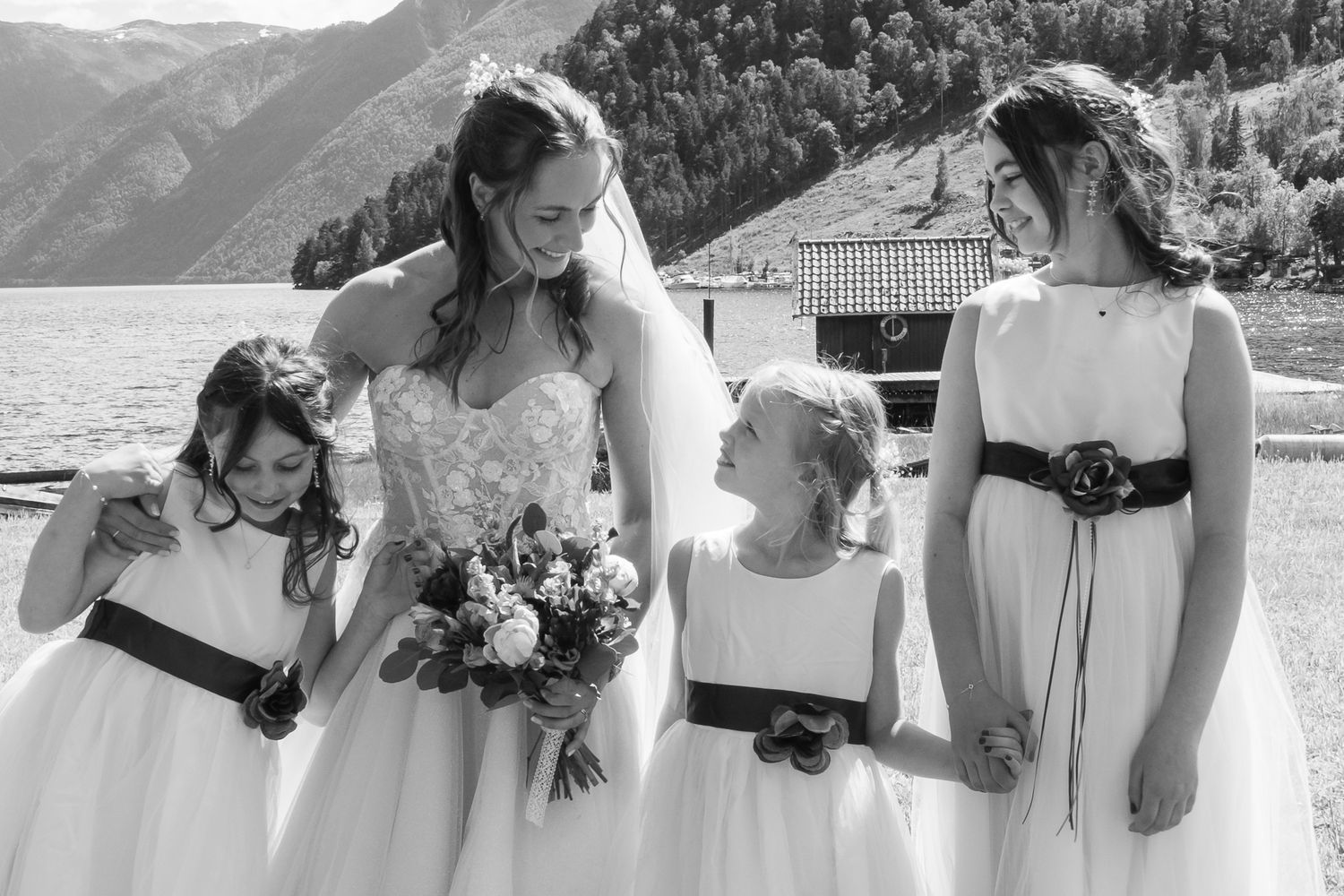 Bride with bouquet and three flower girls in white dresses with black sashes by a mountain lake.