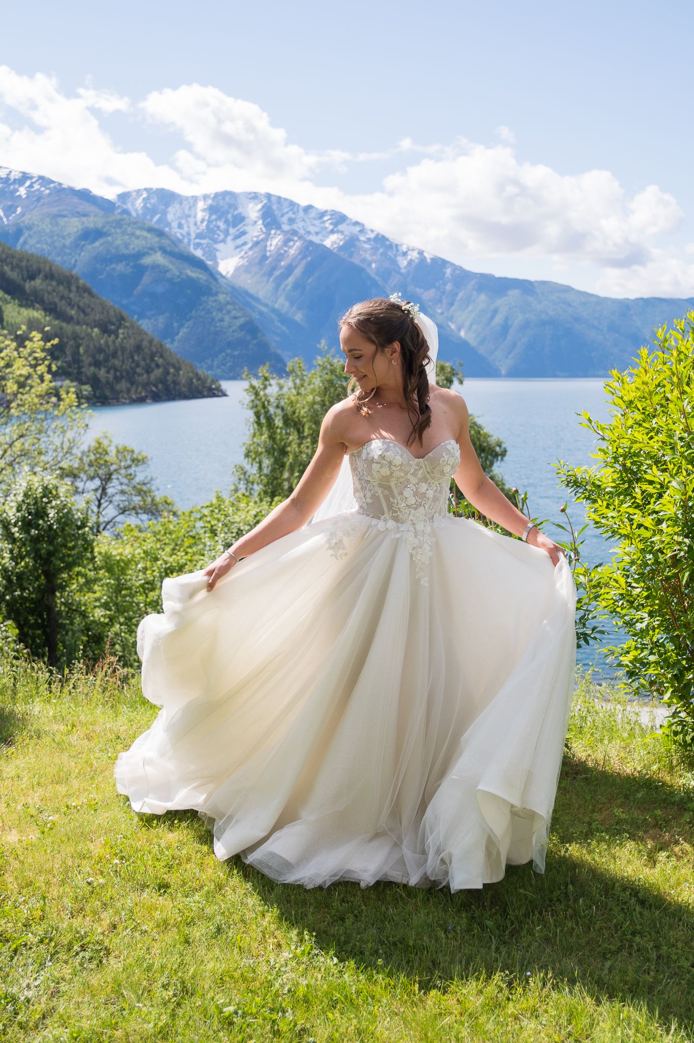 Bride twirls in strapless tulle ball gown on green lawn with snow-capped mountains and blue fjord.