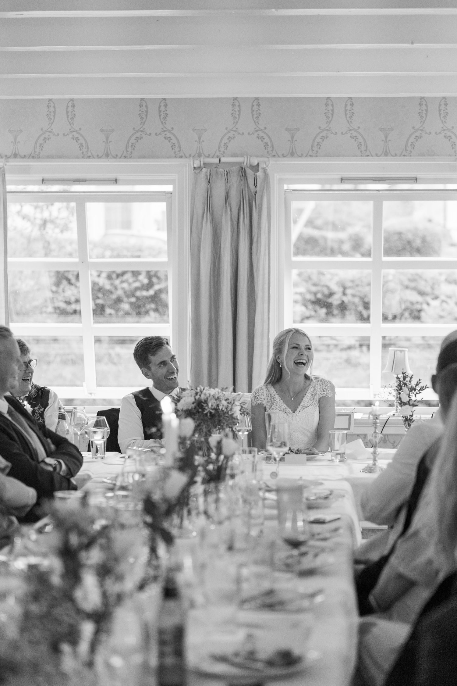 Black and white photo of a laughing bride and groom seated at a wedding reception dinner table with guests.