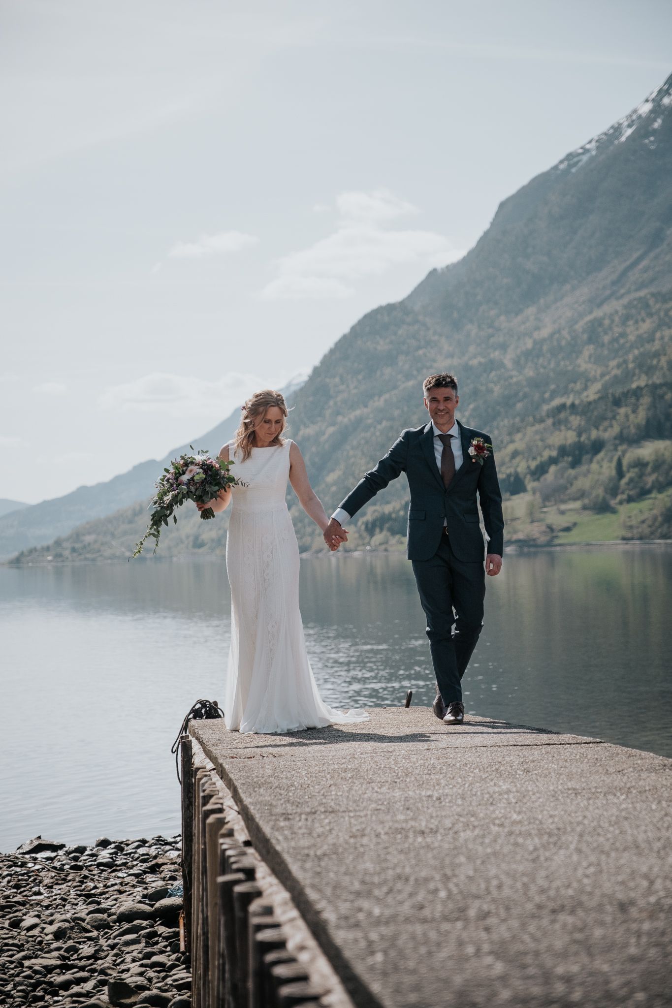 Bride and groom walking hand in hand on a lakeside dock with stunning mountain scenery in the background.