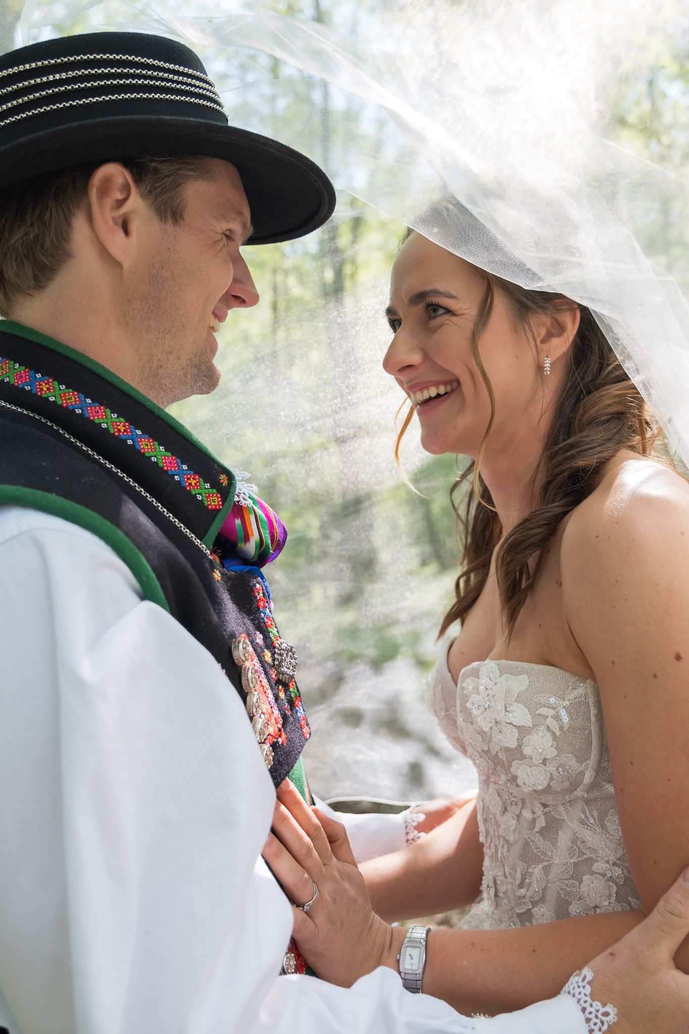 Smiling bride in lace wedding dress gazes at groom in colorful Norwegian bunad under a flowing white veil.
