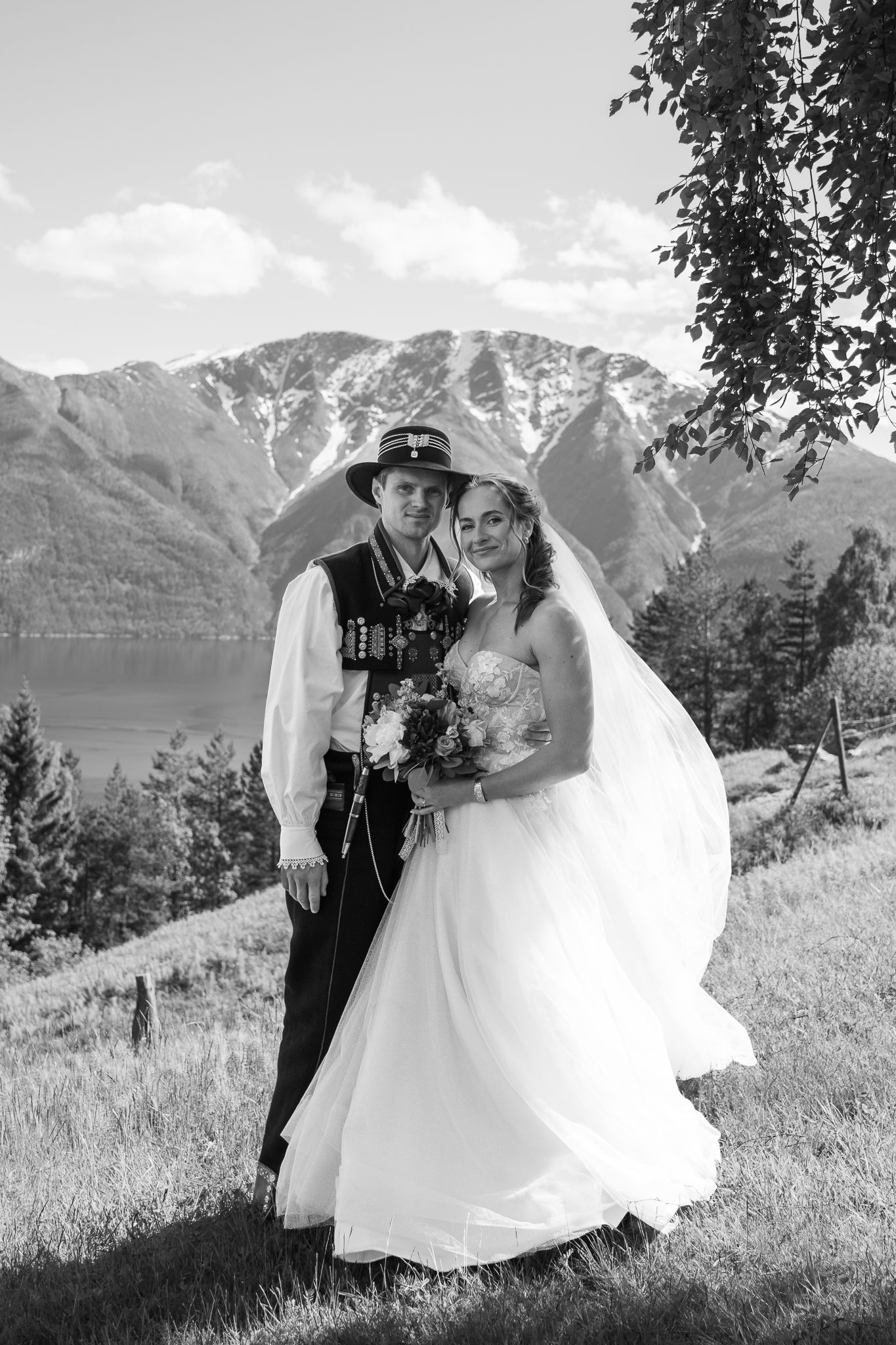 Black and white photo of bride and groom in traditional Norwegian bunad attire posing before a scenic fjord and mountain backdrop.