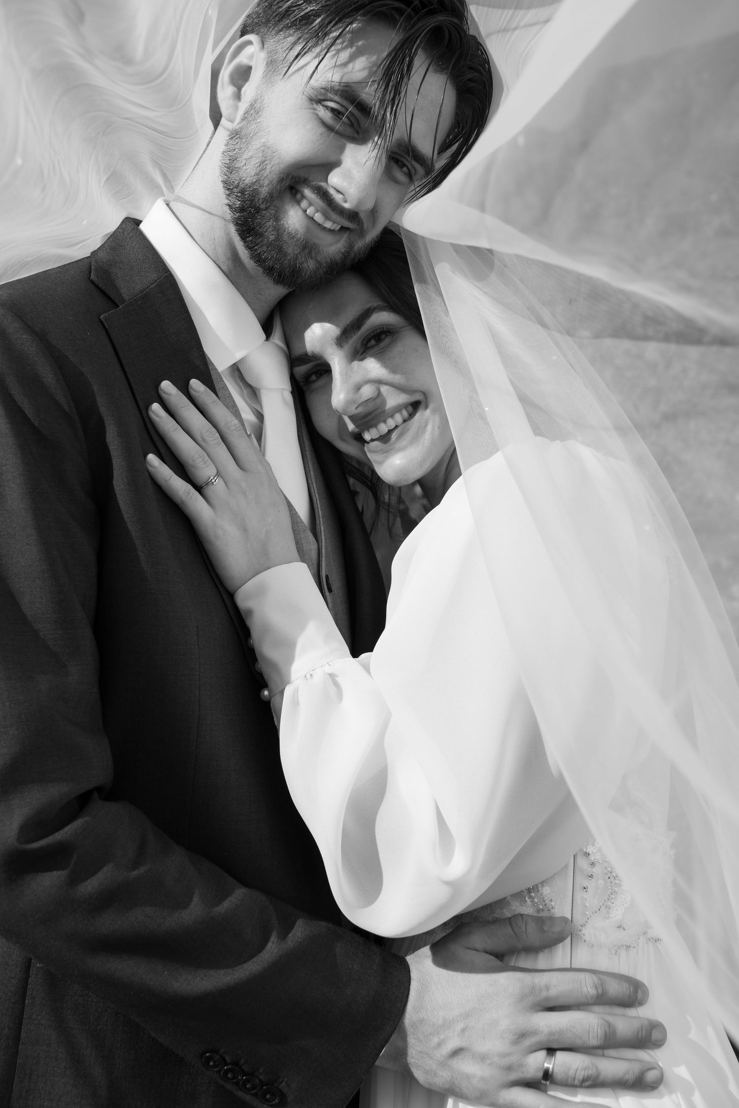 Black and white portrait of a smiling bride and groom embracing under a flowing wedding veil.