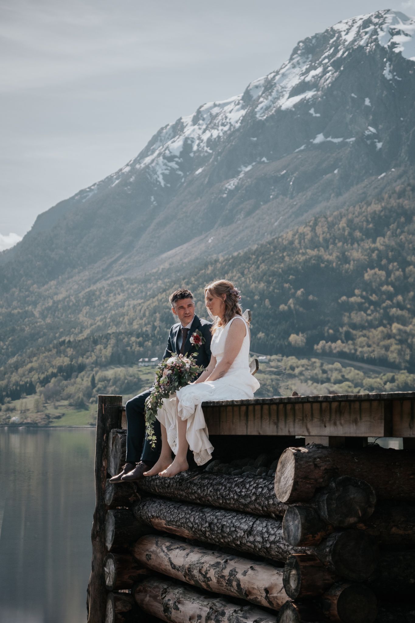 Bride and groom sitting on stacked logs beside a calm lake with snow-capped mountains behind them.