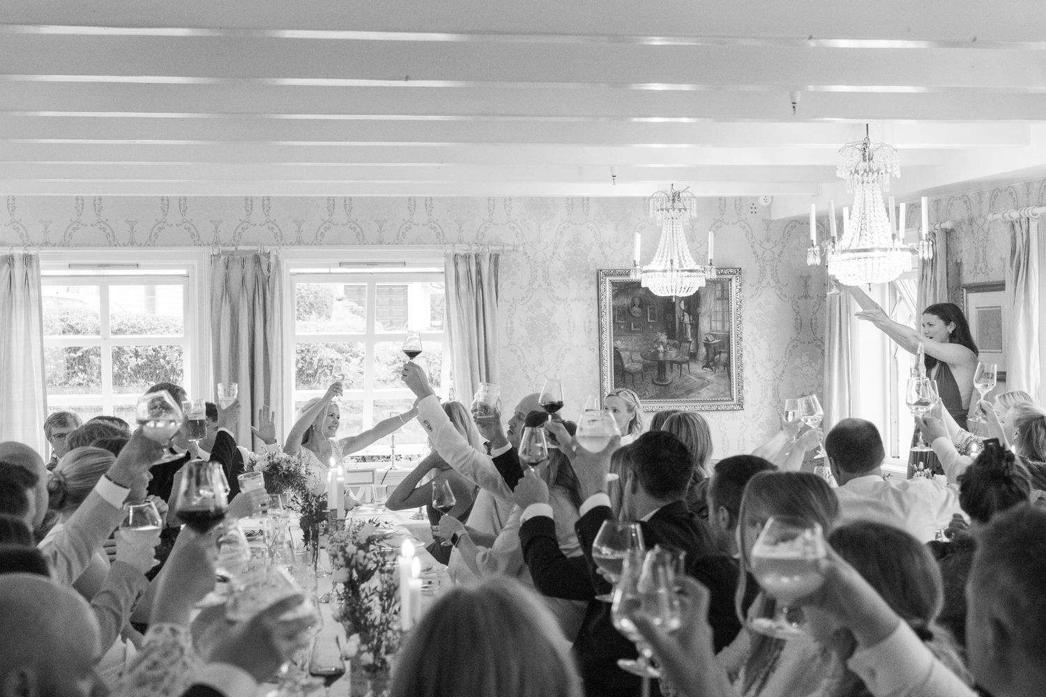 Wedding reception guests raising glasses in a toast inside elegant chandelier-lit dining room.