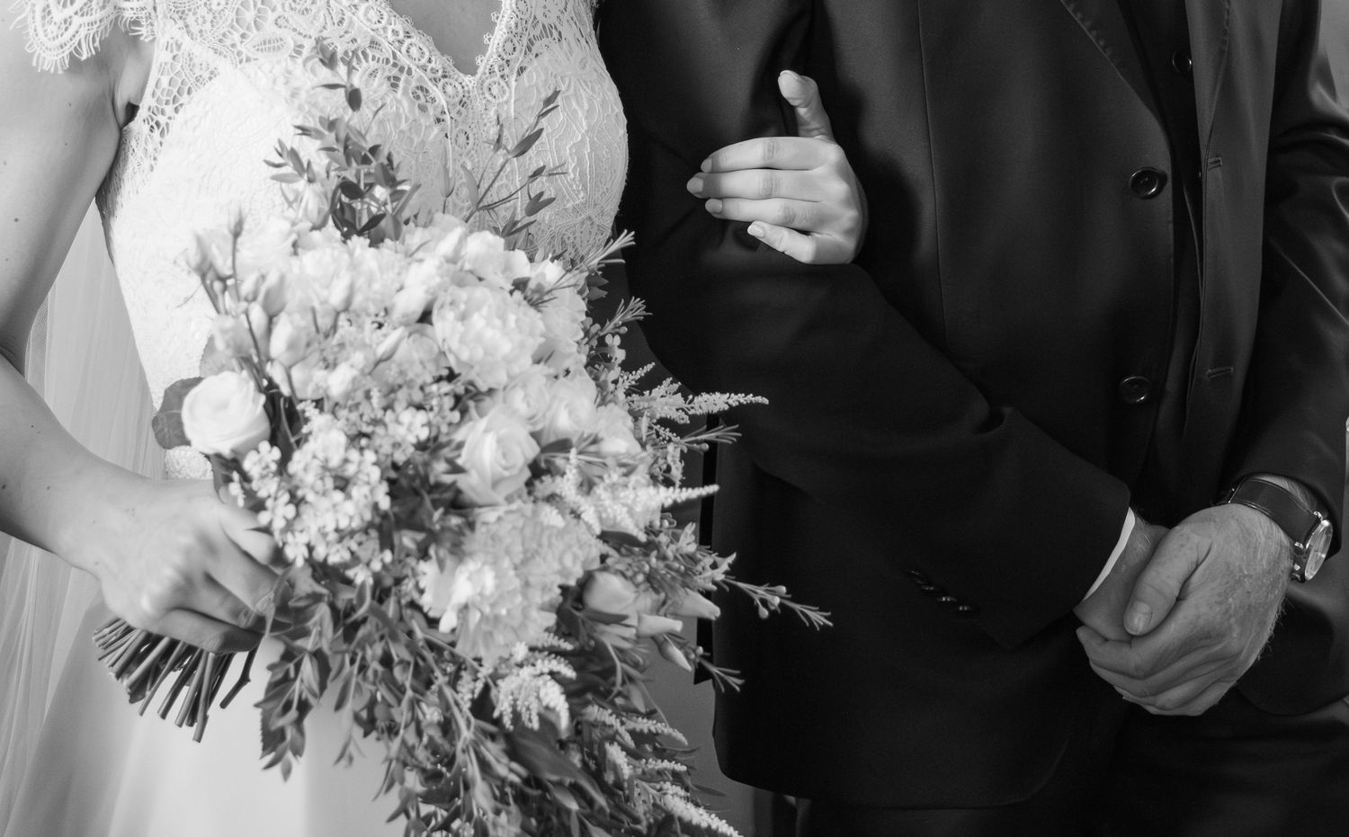 Black and white close-up of bride holding floral bouquet while holding grooms arm in dark suit.