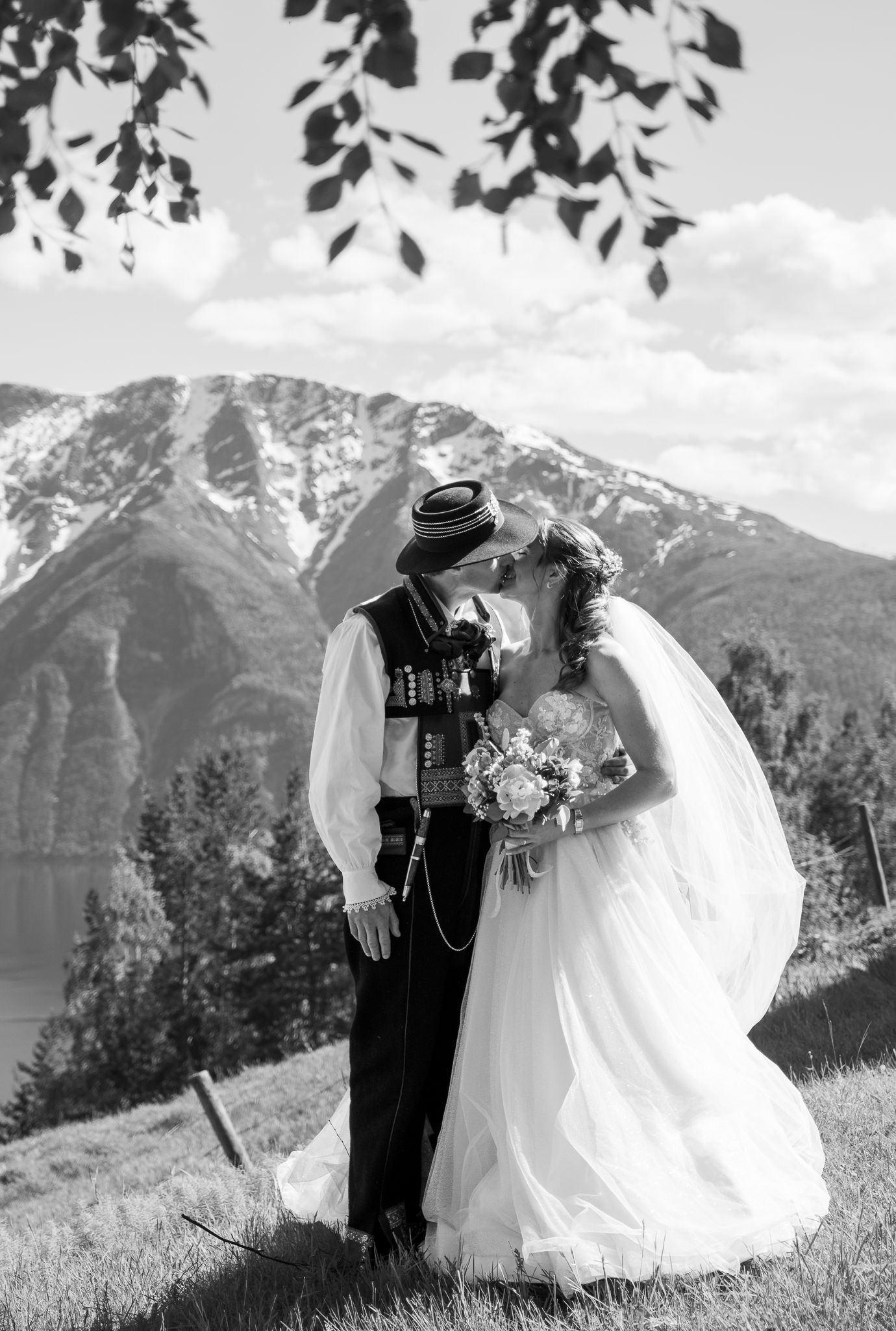 Black and white romantic photo of bride and groom kissing in traditional Norwegian attire with mountain fjord background.