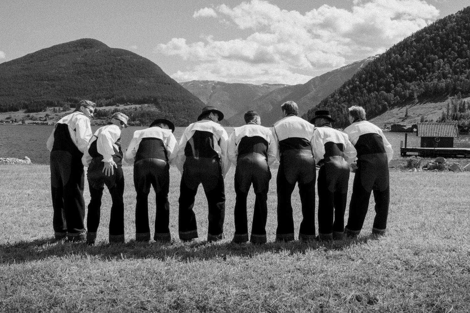 Eight groomsmen in traditional Norwegian bunad attire standing in a row, backs to camera, mountain backdrop.