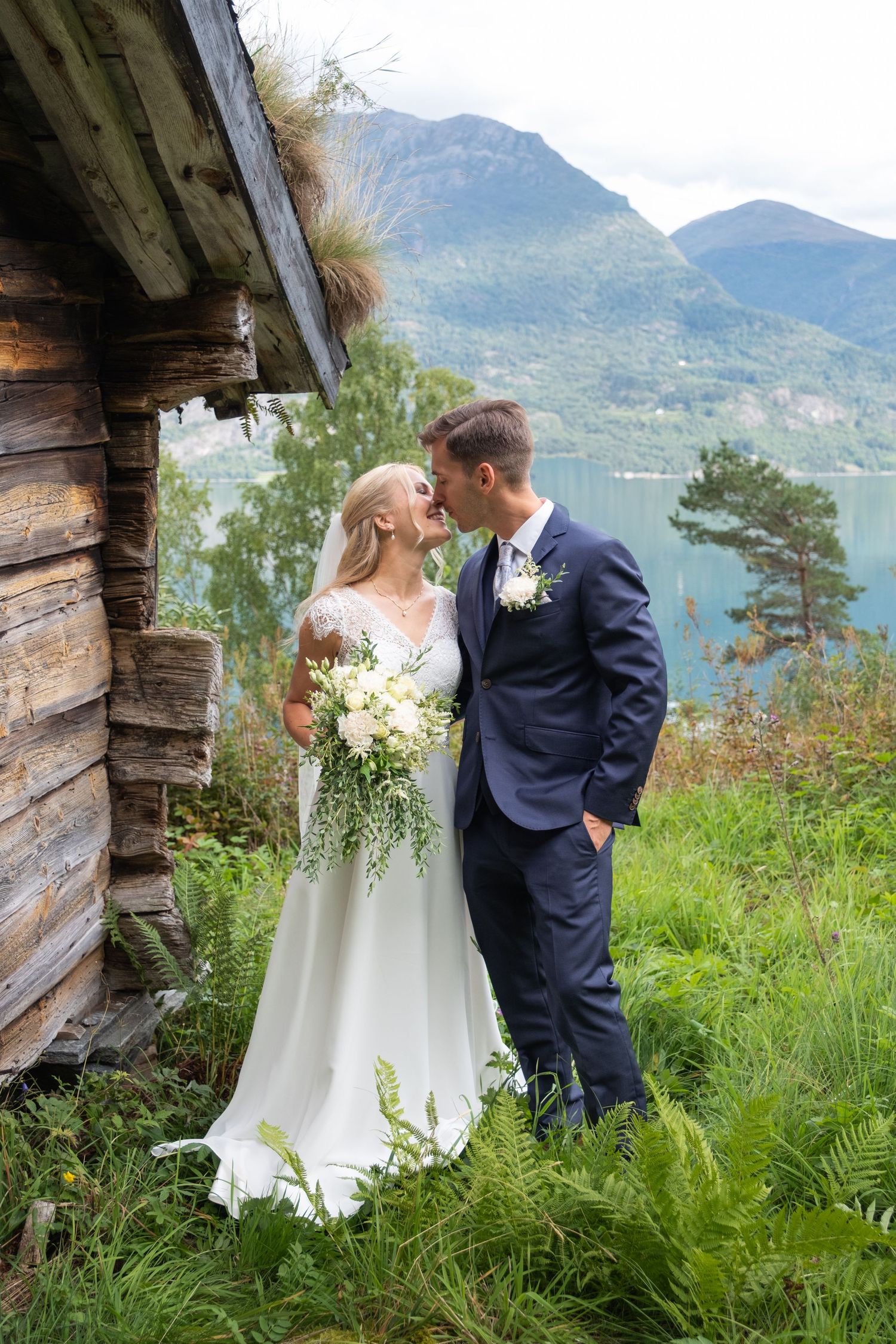 Bride and groom share a romantic kiss near wooden cabin with stunning Norwegian fjord landscape.