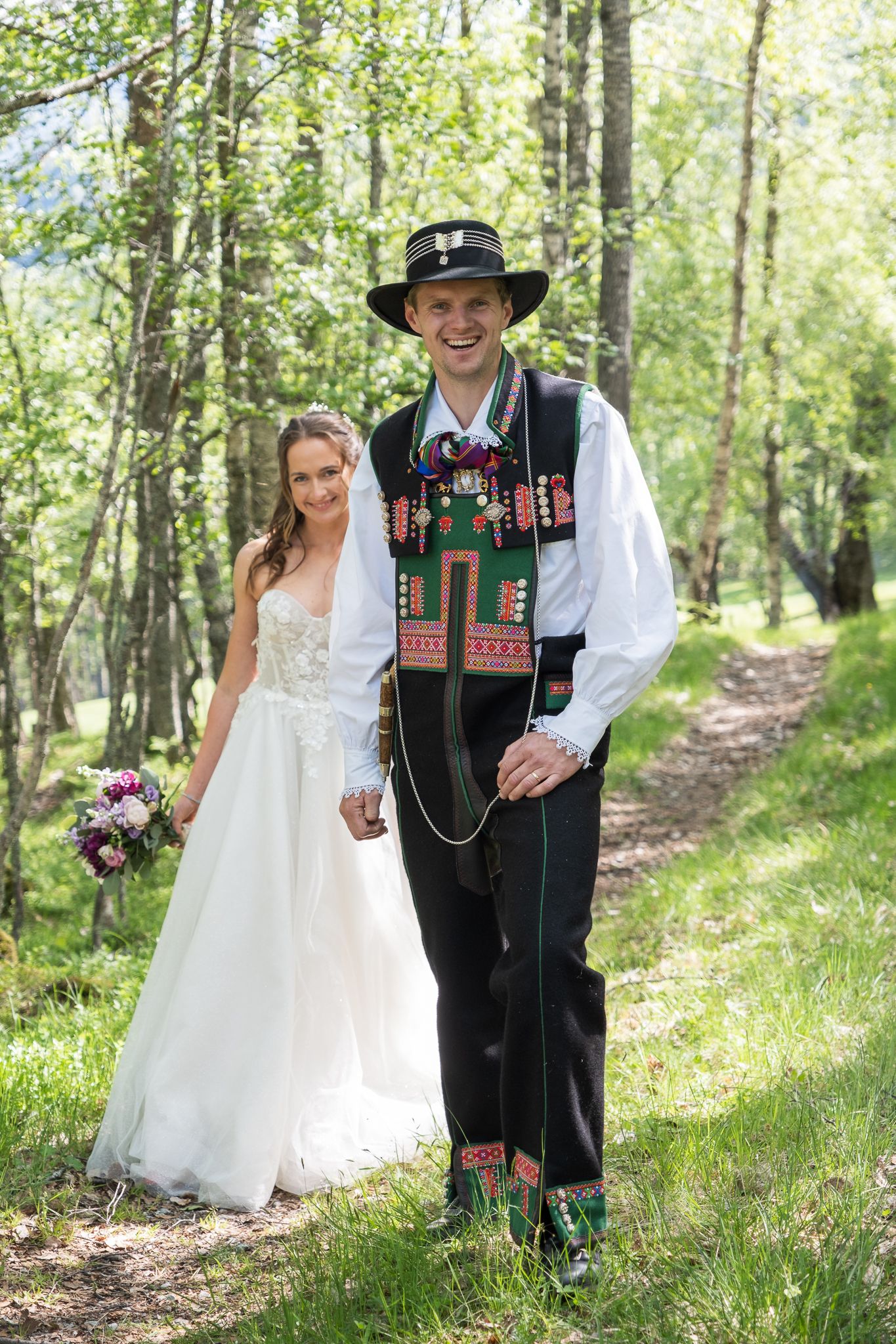 Smiling groom in traditional Norwegian bunad and black hat walks with bride holding a bouquet in a sunny birch forest.